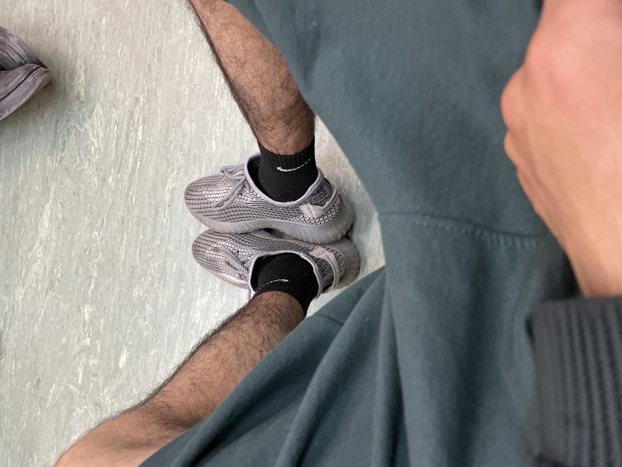 The image is an eye-level, medium shot of a person's legs and feet, seated on a light green, marbled floor. The person is wearing dark green sweatpants and black socks with a white Nike logo. Their feet are clad in grey and white patterned athletic shoes, possibly Yeezy Boost 350s. The person's legs are covered in dark hair, and the scene is well-lit. No specific location in Moscow, Russia, can be determined from the provided image.