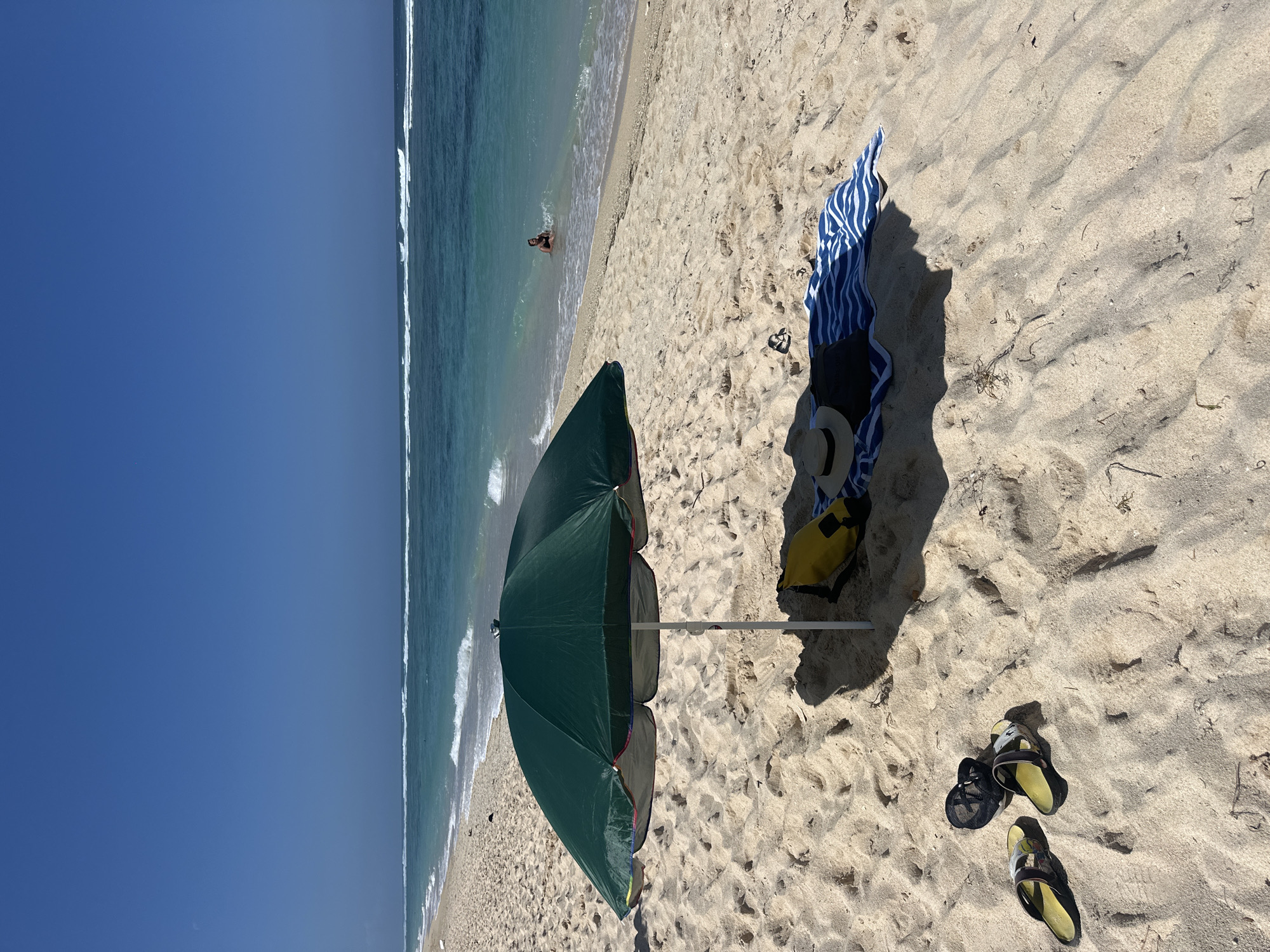 A bright, sunny day unfolds on a sandy beach in Kuta Selatan, Indonesia. The foreground is dominated by the coarse, light-colored sand, marked by footprints and scattered dry vegetation. A large, dark green beach umbrella is positioned centrally, casting a prominent shadow on the sand. Beneath the umbrella, a yellow bag, a straw hat, a dark item of clothing, and a blue and white striped towel are laid out. To the left of the umbrella, a pair of black sandals and a pair of yellow-soled flip-flops rest on the sand. The middle ground features the turquoise ocean, with gentle waves breaking near the shore. In the shallow water, a single individual, appearing to be a person, is partially submerged, facing towards the beach. The background shows the vast expanse of the ocean meeting a clear, deep blue sky.