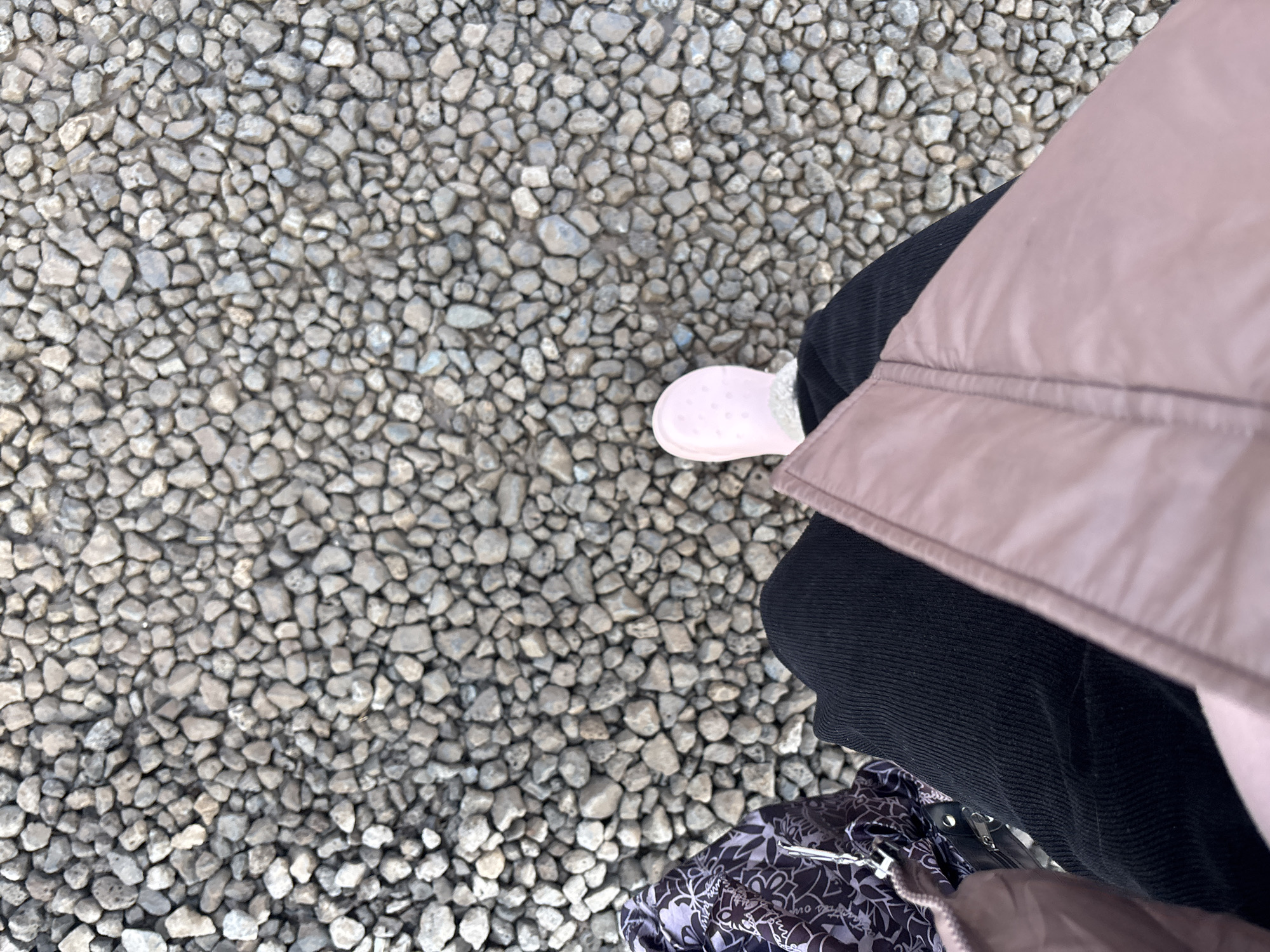 The image shows a view from above, focusing on the lower portion of a person standing on a surface covered with small, light-gray stones. The person is wearing dark corduroy pants and a light-pink puffer jacket. A pink slip-on shoe is visible on the left side of the frame. The bottom of a black and white patterned bag is partially visible in the lower left corner. It is not possible to identify the location of the city of Ferma, Russia.