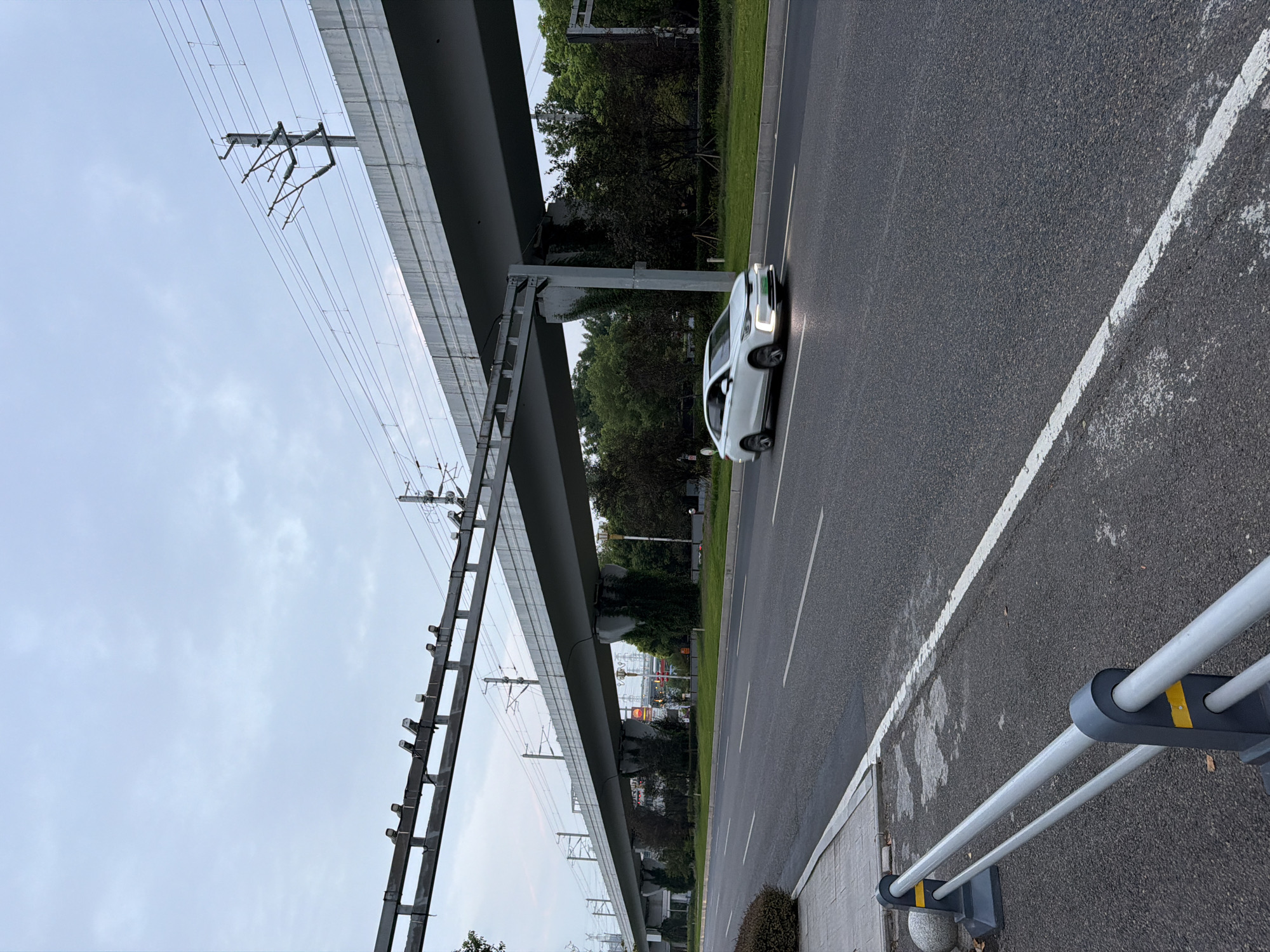 The image depicts a road in the city of Jiangjin, China, with an elevated railway track overhead. The sky is overcast. A white sedan is traveling on the road, its headlights illuminated. The road has white lane markings. To the left of the road is a sidewalk with a decorative bollard and a metal barrier. Behind the road and sidewalk, there is a strip of green grass and trees, with the concrete pillars supporting the elevated railway track visible among the trees. The elevated railway structure itself is a prominent feature, with overhead electrical lines and supports.
