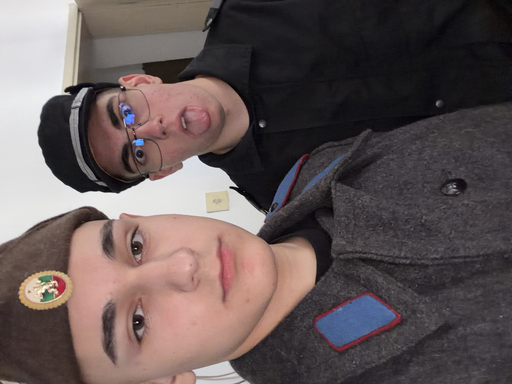 Two young men pose for a selfie. The man in the foreground wears a gray-colored military jacket with a blue and red patch and a matching hat with a gold emblem depicting a lion and other symbols. The second man wears a black military jacket and a black cap with silver trim, and he has glasses and is sticking out his tongue. A light switch is visible on the wall behind them. The location appears to be indoors in an unknown location in Panagyurishte, Bulgaria.
