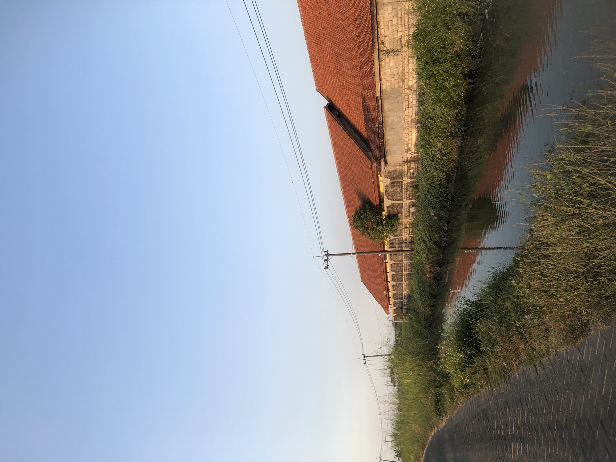 The image depicts a scene in Keboguyang, Indonesia, featuring a paved path on the left, bordered by tall grass and vegetation. Adjacent to the vegetation is a narrow canal with calm water reflecting the sky and the buildings. On the right side of the canal, a long, low building with an orange tile roof stretches horizontally across the frame. The building is constructed of light-colored bricks with a concrete foundation or wall along its base. Several utility poles with power lines are visible along the length of the building and in the distance. The sky is a clear, pale blue, indicating daylight, likely late afternoon or early morning given the soft light. The overall atmosphere is peaceful and rural. There are no people or animals visible in the image.