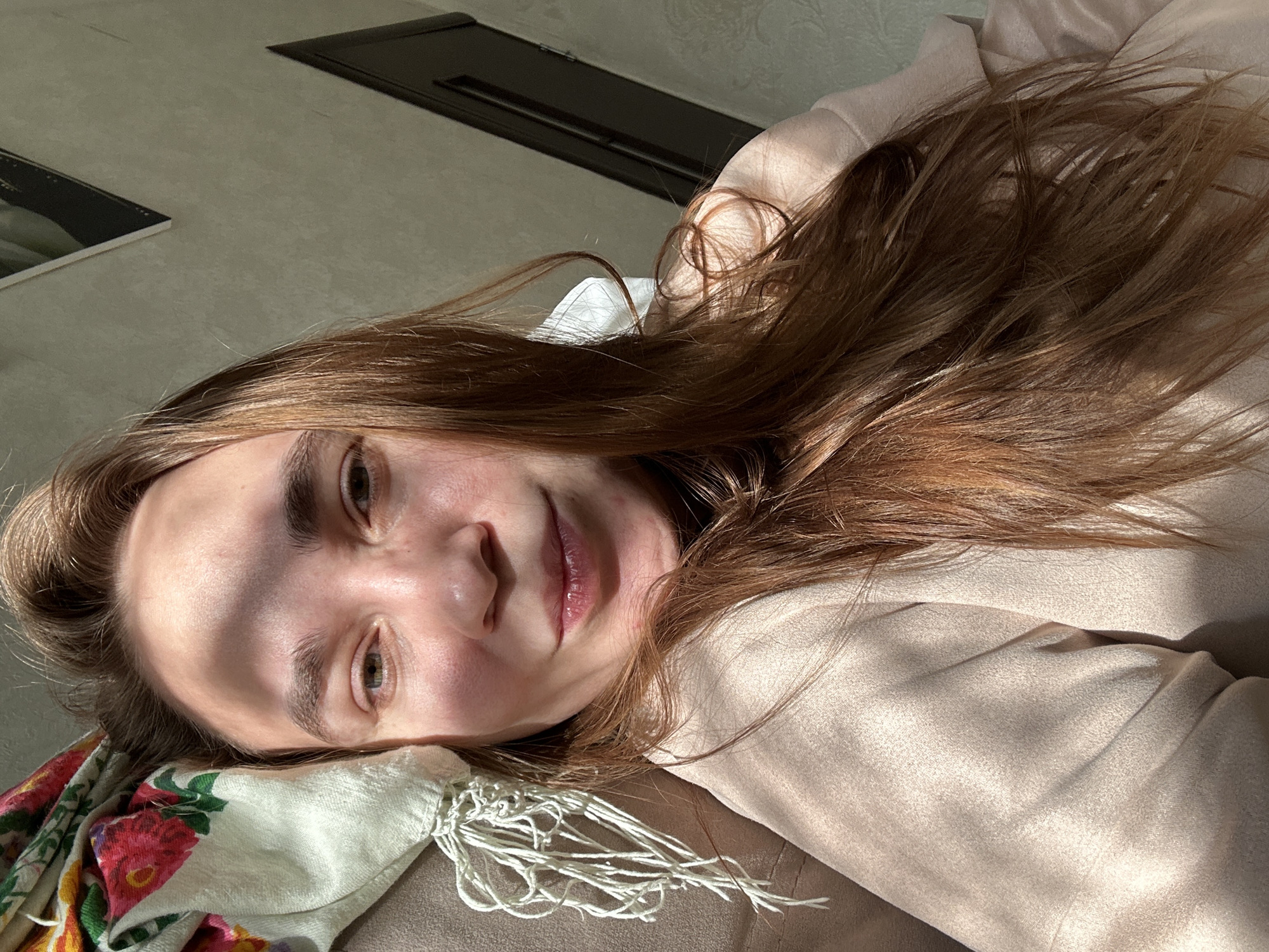 A young woman is captured in a close-up, tilted shot, lying down indoors in Uva, Russia. Her head and upper torso are visible as she looks directly towards the camera with a calm, slightly smiling expression. Her long, reddish-brown hair is spread around her head and shoulders.
She rests on a plain, light brown or beige surface, which appears to be a sofa or bed. A floral-patterned fabric, possibly a scarf or blanket, with red/pink flowers, green leaves, and white fringes, is visible on the left side of the frame, underneath her head. The woman is wearing a light beige or cream-colored top, which appears to be made of a soft material.
Strong, direct sunlight illuminates her face and hair, creating distinct shadows across her forehead, nose, and cheek, indicating it is daytime, likely morning or late afternoon. The lighting gives the scene a warm, natural ambiance. In the background, parts of a light-colored wall and two dark, rectangular objects (possibly shelves or architectural features) are visible, suggesting a home setting. There is no visible text in the image.