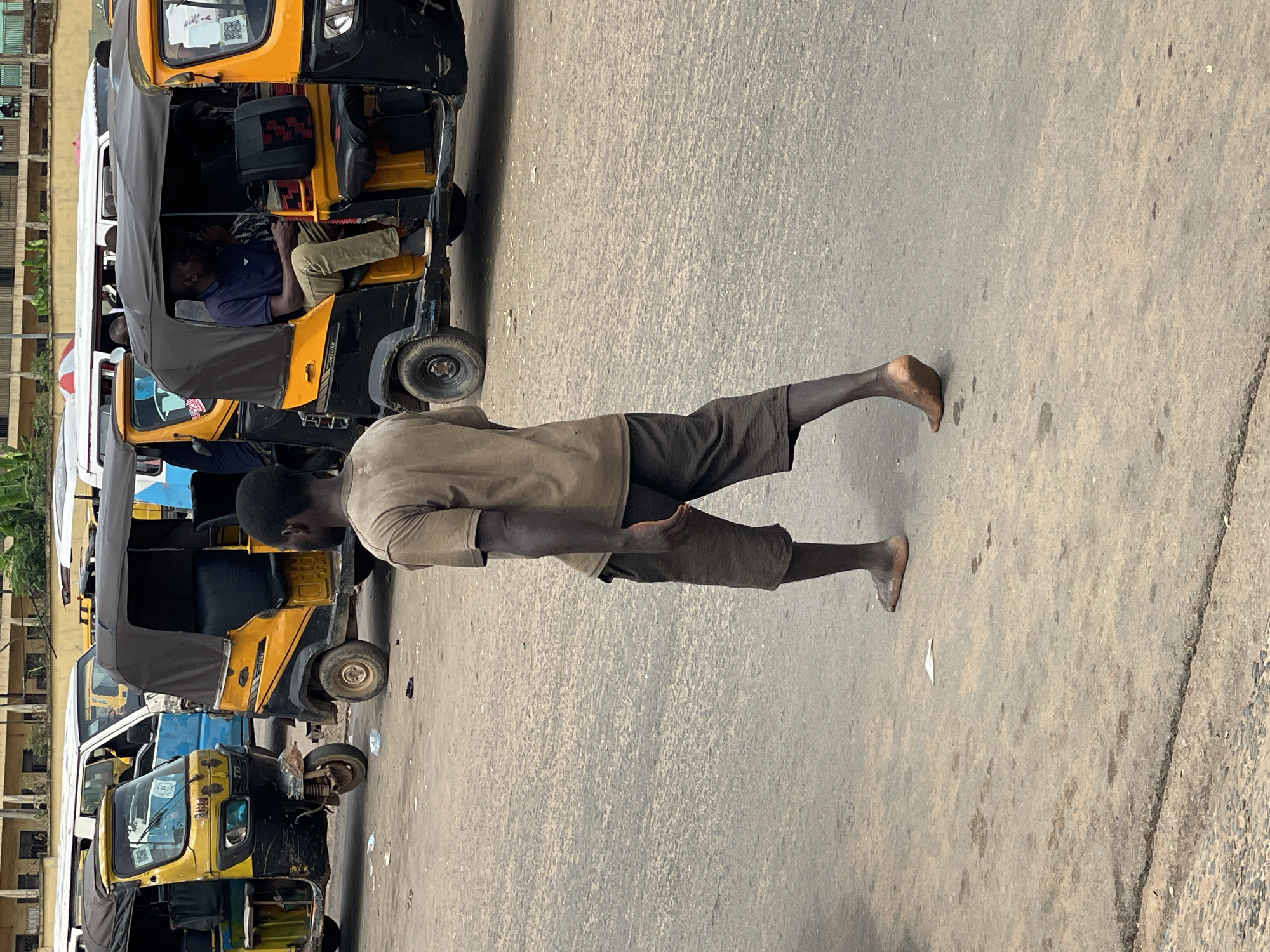 A medium shot shows a man walking barefoot on a paved street in Onitsha, Nigeria. The man has dark skin and short hair. He is wearing a short-sleeved khaki shirt and knee-length brown shorts. He is walking away from the viewer, and his arms hang at his sides.
Parked along the left side of the street are several auto rickshaws. Most are yellow with black trim and white canvas tops. People are sitting inside some of the rickshaws. A multistory building appears in the background. The sky is overcast.