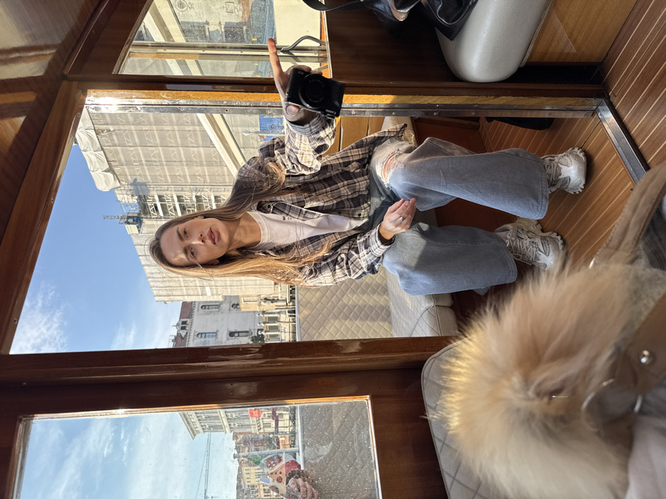 A young adult female is seated inside a wooden watercraft, likely a water taxi, on a canal in Venice, Italy, under a clear blue sky. She wears a plaid shirt over a white t-shirt, ripped light blue baggy jeans, and white chunky athletic shoes. Her long brown hair is down, and she looks directly at the camera with a neutral expression. Her right arm is extended outwards, holding a black compact digital camera and pointing towards the right side of the frame. A silver ring is visible on her left ring finger. The boat's interior features polished wooden walls and floor, with white diamond-stitched upholstered seating. A blurred, fluffy, light-colored object is visible in the immediate foreground, positioned on a seat. A black bag rests on the seat to the woman's right. Through the boat's windows, typical Venetian architecture is visible along the canal. In the background, a multi-story building is largely covered in white construction scaffolding, with a blue crane visible at its top, indicating ongoing construction or renovation. The scene takes place during daylight hours.