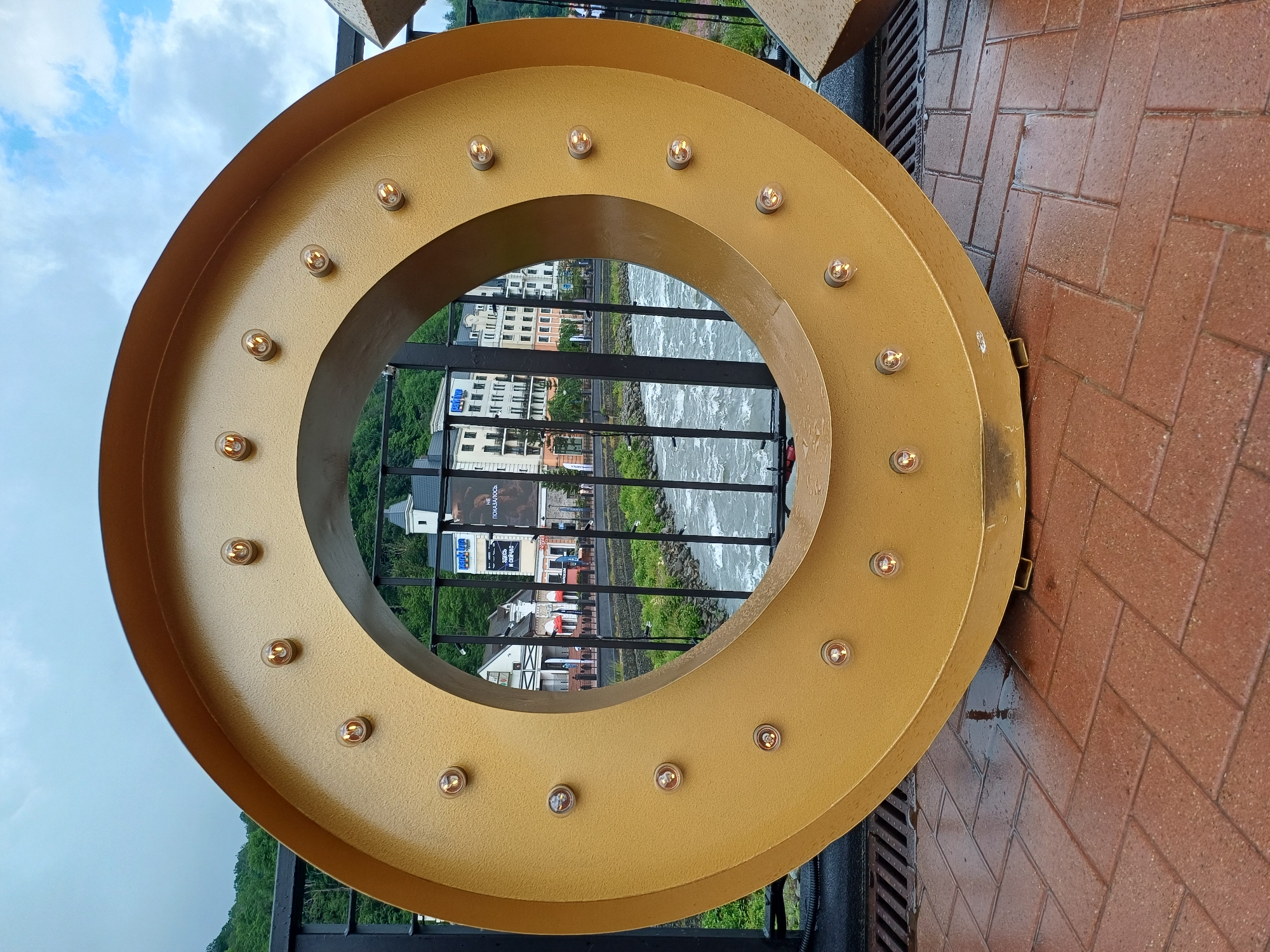 The image shows a large, circular, gold-colored decorative object with lights, positioned outdoors on a brick pathway. Behind the object, a black metal railing separates it from a river with turbulent water. Across the river, a row of buildings with light-colored facades are visible, nestled amongst green trees. The sky is cloudy. The scene appears to be in the city of Estosadok, Russia, as indicated by the architecture and natural surroundings typical of a resort town. The pathway is wet, suggesting recent rain. There are no people clearly visible in the frame. The decorative object itself is the primary subject, with the background providing context for its location.