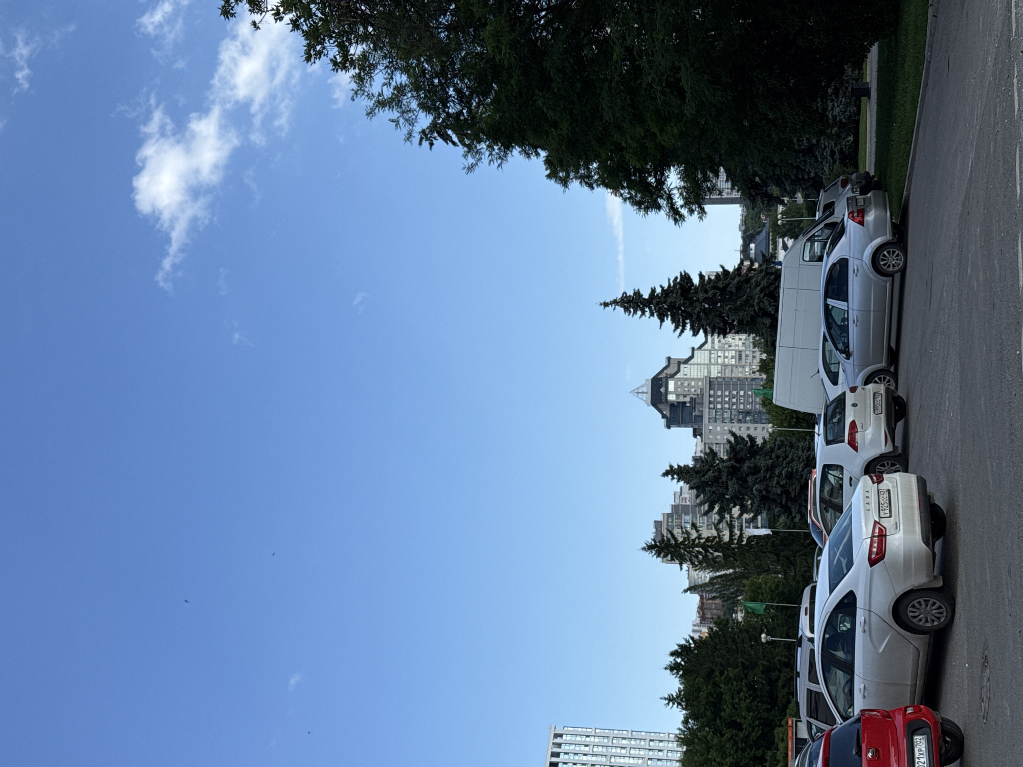 The image depicts a parking lot in a city under a clear blue sky with scattered white clouds. In the foreground, several cars are parked, including a red Kia on the left, a white Lada, a white Volkswagen Golf, and a silver Chevrolet. The cars are arranged in rows on an asphalt surface. Behind the parking lot, a line of evergreen trees, including several tall pine trees, is visible. In the background, there are modern apartment buildings of varying heights, some with distinctive architectural features. The overall scene suggests a typical urban environment on a clear day.