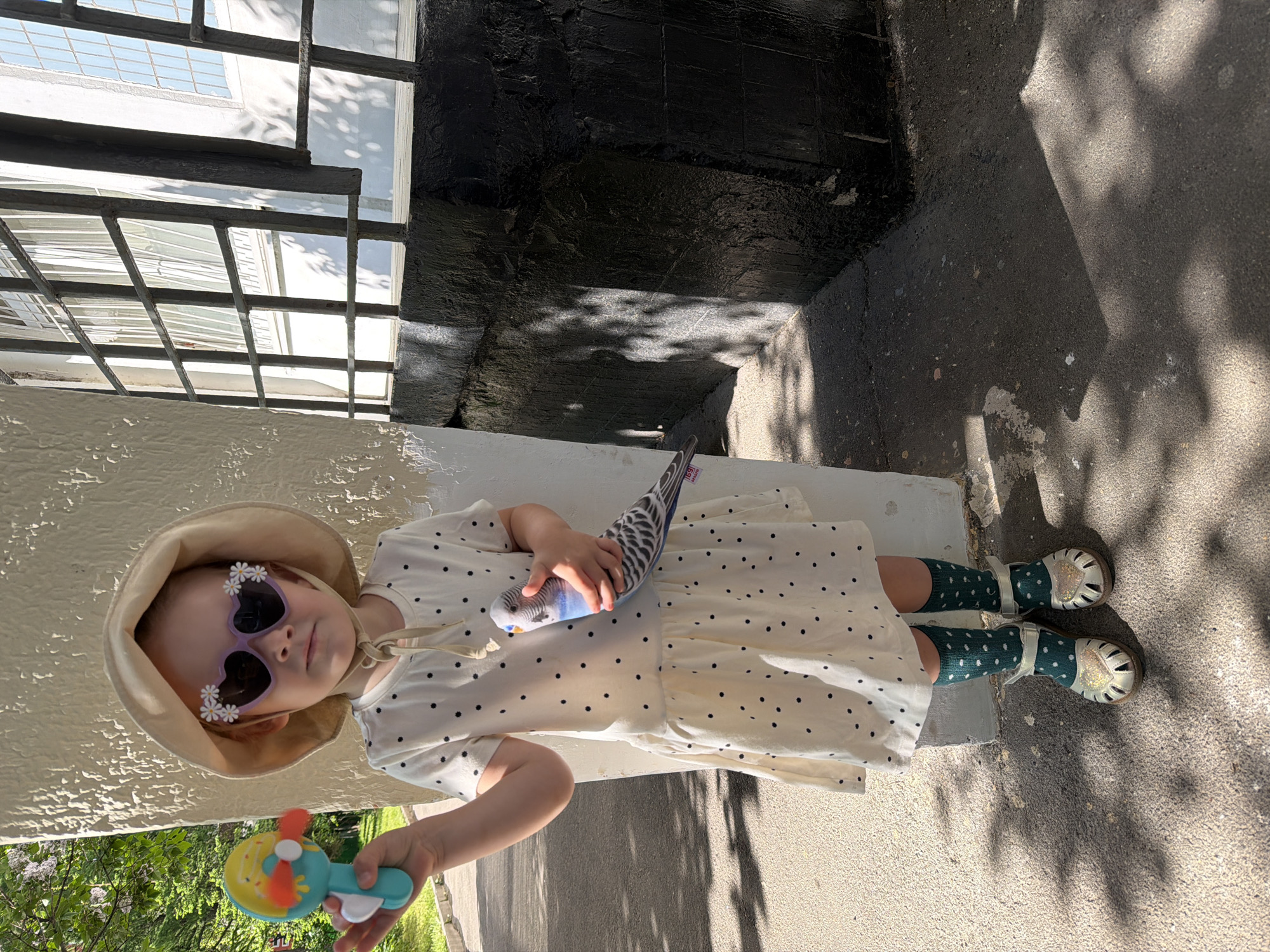 A young child stands in an exterior urban setting. The child is wearing a light-colored dress with black polka dots, green polka-dot socks, silver sandals, a light-colored sun hat, and sunglasses with flower embellishments. The child is holding a toy in one hand and a parakeet in the other. The setting includes a building with a black and white facade and window grilles, and a concrete surface with shadows cast by foliage. The location cannot be definitively identified as Moscow, Russia from the visual information provided.