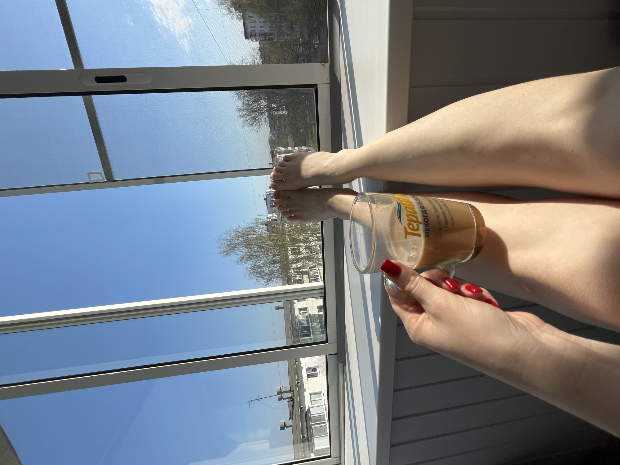 The image shows a partial view of a person sitting by a window. The lower part of the legs and feet are visible, with a cup of coffee or tea held in their hand. The person has red nail polish. The window looks out onto a city scene with buildings and trees, under a clear blue sky. The location is Chaikovsky, Russia, based on the "Teprada" brand writing on the glass, which appears to be a locally made brand of coffee. The window frame appears to be white. The overall scene suggests a relaxed setting, possibly a sunny morning or afternoon.