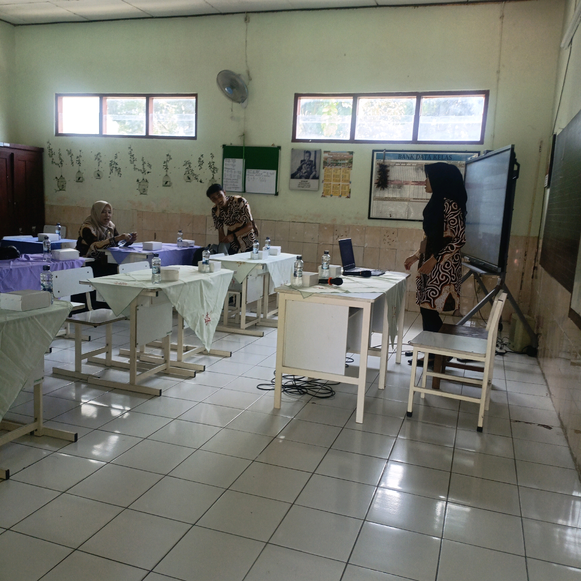 A classroom or meeting room in Salatiga, Indonesia, is depicted. The room has light green walls and a tiled floor. Several tables are arranged, some covered with colored cloths, holding water bottles and boxes. In the foreground, a white chair is positioned near a table. Two individuals are visible: a woman in a headscarf and patterned dress is seated at a table on the left, and another woman in a similar headscarf and patterned dress stands near a large screen on the right. A man in a patterned shirt is also present, standing between the tables. Windows are visible along the walls, and some posters are displayed. The overall impression is of an indoor gathering or instructional setting.