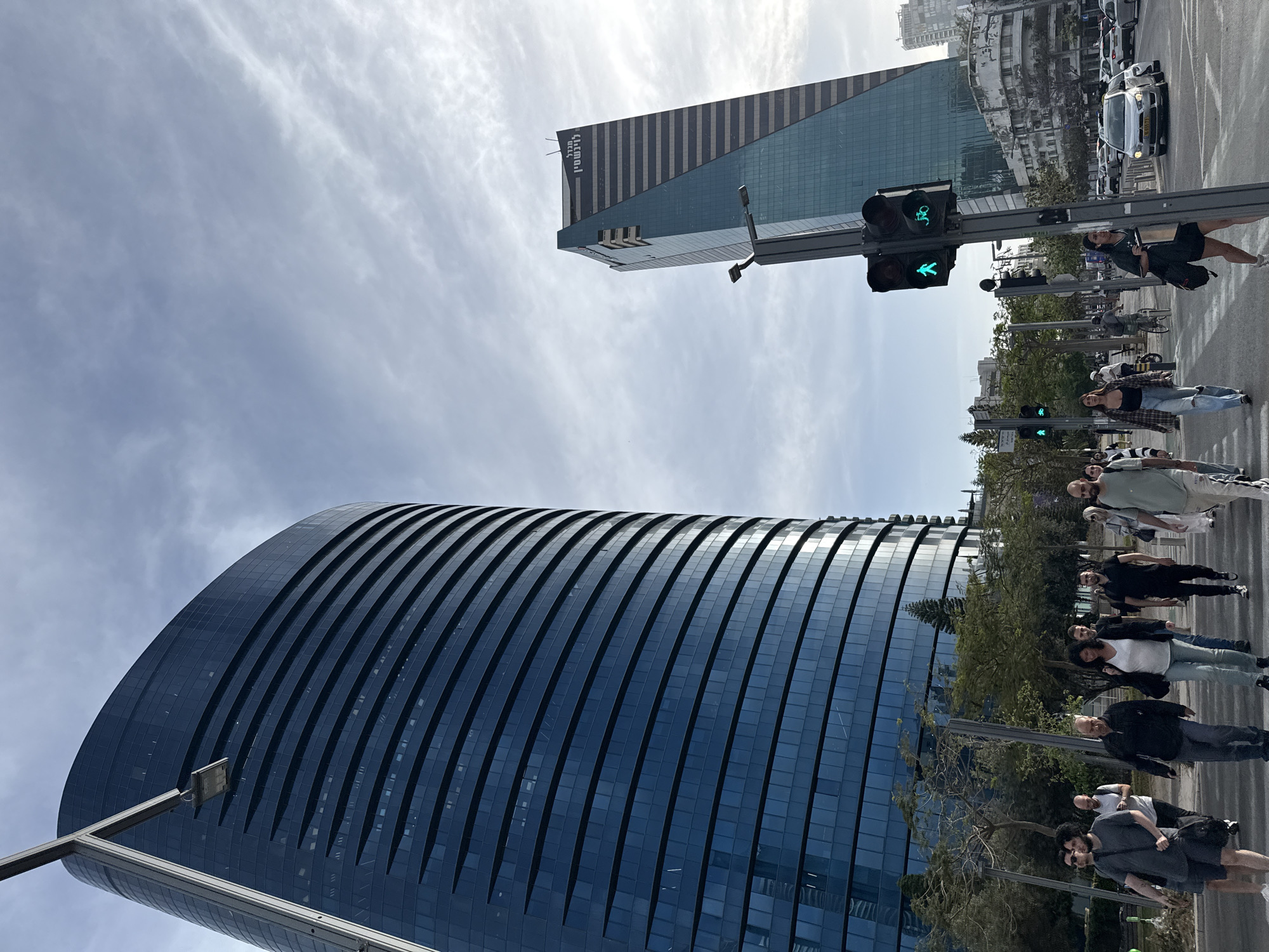 The image shows a street scene in Tel Aviv, Israel, with pedestrians crossing a street at a crosswalk. The traffic light shows a green "walk" signal. Two modern skyscrapers are visible in the background. One is a curved, blue glass building. The other is a taller, angular building with a dark stripe on its upper section. Cars are lined up on the street adjacent to the crosswalk. The sky is partly cloudy.