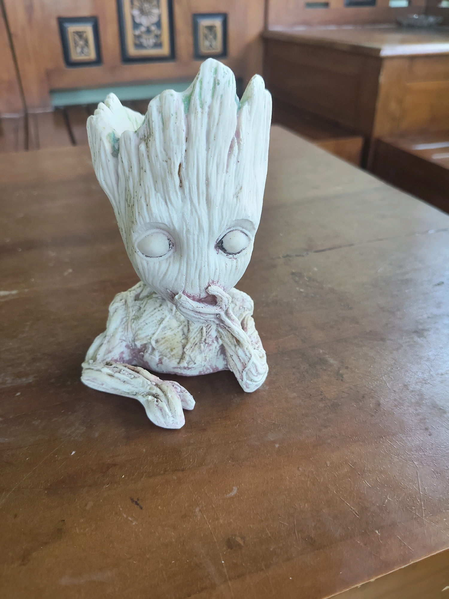 A small, white, ceramic figurine of Groot from Guardians of the Galaxy sits on a polished wooden table. The figurine is in a seated position with its hands near its face, and it has a slightly distressed, textured finish with hints of pink and green. The wooden table surface is worn and shows visible grain and scratches. In the background, out of focus, are framed pictures on a wooden wall and the edge of wooden furniture, suggesting an indoor setting. The overall lighting is soft, and the scene appears to be a still life composition.