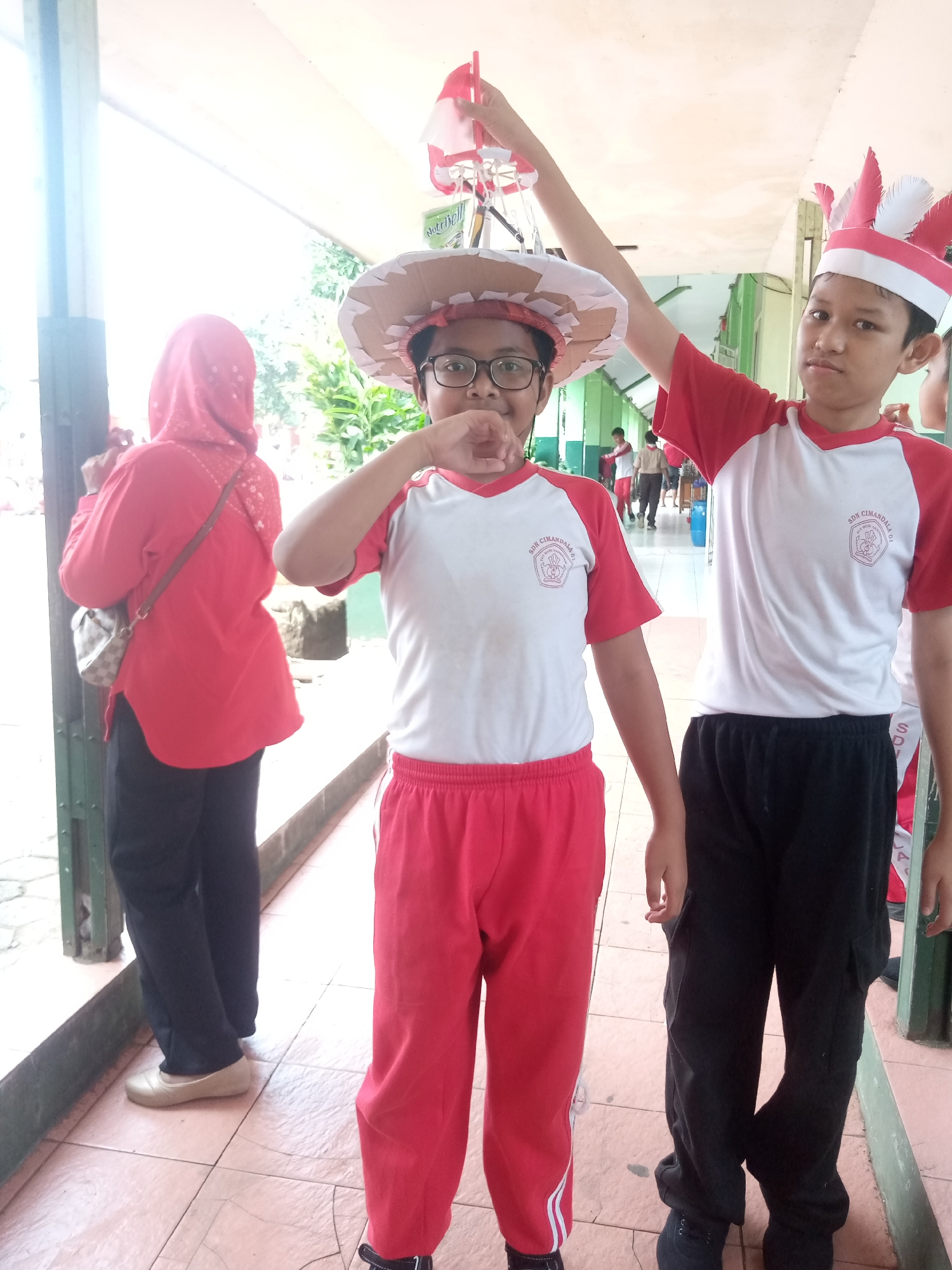Two young boys stand in a school hallway in Sukaraja, Indonesia, dressed in red and white athletic attire. The boy on the left wears a wide-brimmed hat made of cardboard and paper, possibly part of a costume or school project. He is also wearing glasses and a red t-shirt with a white panel featuring a school logo. The boy on the right wears a red and white headband with feather-like decorations, a red and white t-shirt with the same school logo, and black pants. He holds his right arm up, as if posing. In the background, another person wearing a red hijab and a red shirt is visible, along with other students in the hallway. The hallway has tiled floors and green pillars. The overall scene suggests a school event or celebration, possibly related to Indonesian Independence Day, given the red and white motif.