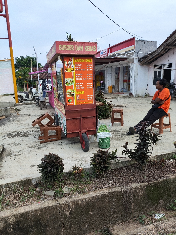 This outdoor scene in Kecamatan Selagai Lingga, Indonesia, features a vibrant red and pink mobile food cart as its central subject. The cart is prominently labeled "BURGER DAN KEBAB" and displays a menu for "MADINAH KEBAB," listing items such as "KEBAB KEJU," "KEBAB TELUR," and various "BURGER" options including "BURGER KEJU," "BURGER TELUR," and "SPESIAL TELUR," all with prices indicated in Indonesian Rupiah. A small graphic with the word "Lezzet" is also visible on the menu.
To the right of the food cart, a man wearing an orange t-shirt and dark pants sits casually on a small brown stool, leaning against a building. Several other identical plastic stools are scattered around the cart, some upright, others overturned. A green plastic basket containing leafy vegetables sits on the ground near the cart's wheel. The foreground features a concrete curb and small decorative plants.
In the background, a red and white Alfamart convenience store is visible, along with another simple white building with a wooden roof structure. Scooters or motorcycles are parked in the distance. The sky is overcast, indicating a cloudy day or soft lighting. Overhead power lines crisscross the upper part of the frame. The scene depicts a quiet moment at a roadside food stall.