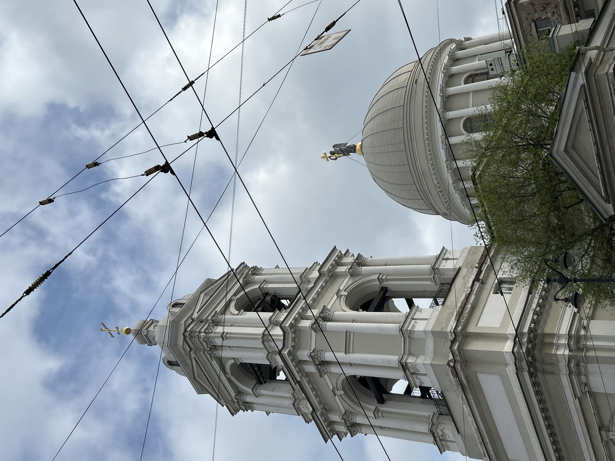 The image is a low-angle view of architectural landmarks in Saint Petersburg, Russia. A bell tower with a golden cross at its peak is on the left, and a dome topped with a golden statue is on the right. Both structures are white and detailed with classical architectural elements. Overhead power lines crisscross the sky, obstructing the view of the partly cloudy sky. Green foliage from a tree is visible to the right of the dome.