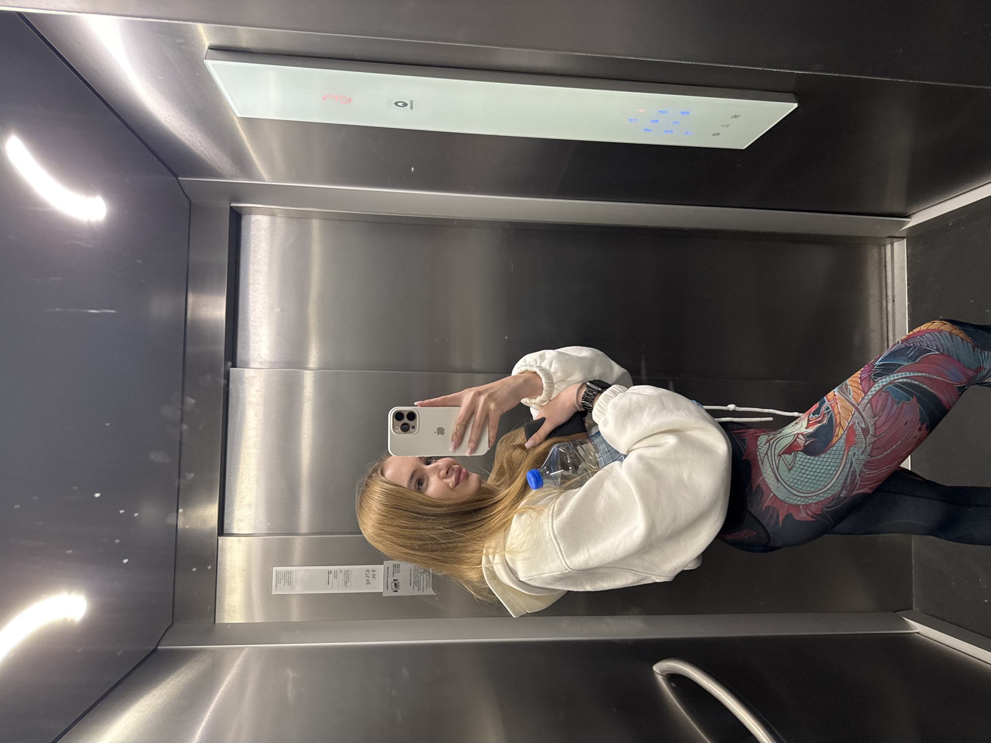 A young woman stands in an elevator taking a selfie with her smartphone. She has long blonde hair, wears a white hooded sweatshirt, patterned leggings, and a watch on her left wrist. She holds a water bottle in one hand and her phone in the other. The elevator has a stainless steel interior with a control panel on the upper part of the back wall. A metal handrail is visible in the lower part of the frame. The location is inferred to be Budva, Montenegro, based on the image prompt.