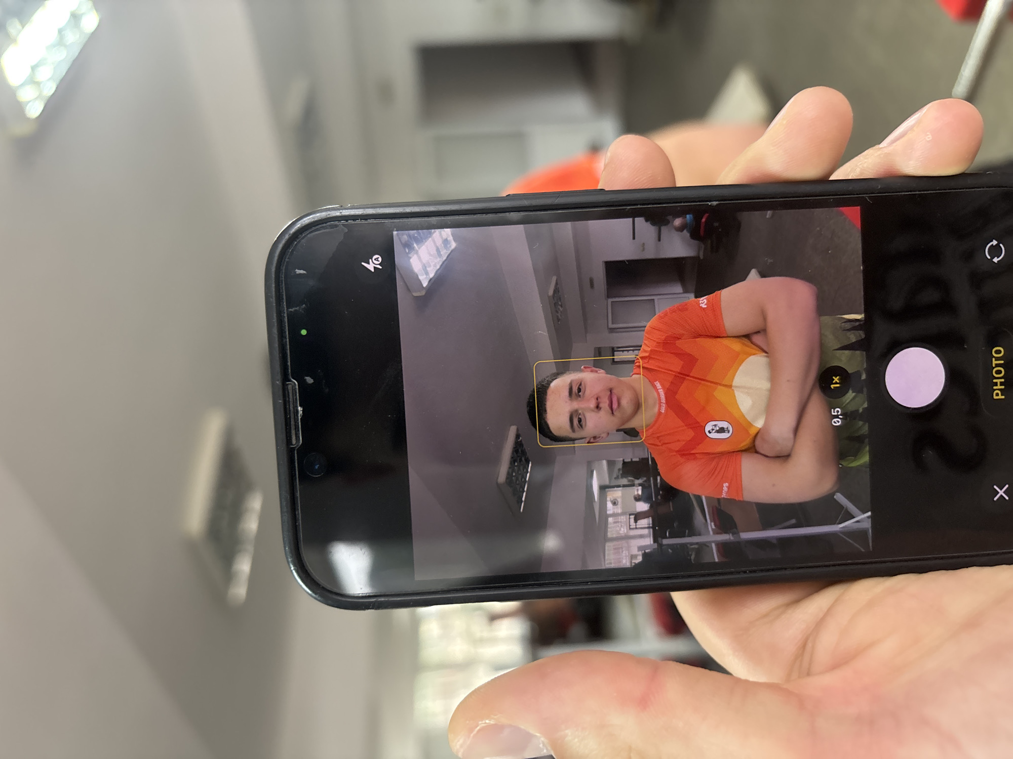 A person is holding an iPhone displaying the phone's camera interface. The camera is focused on a young man in an orange shirt standing with his arms crossed. The background appears to be a gym setting, with weight machines and other gym equipment visible. The location details, "Zlatograd, Bulgaria," cannot be confirmed or denied from the image alone.
