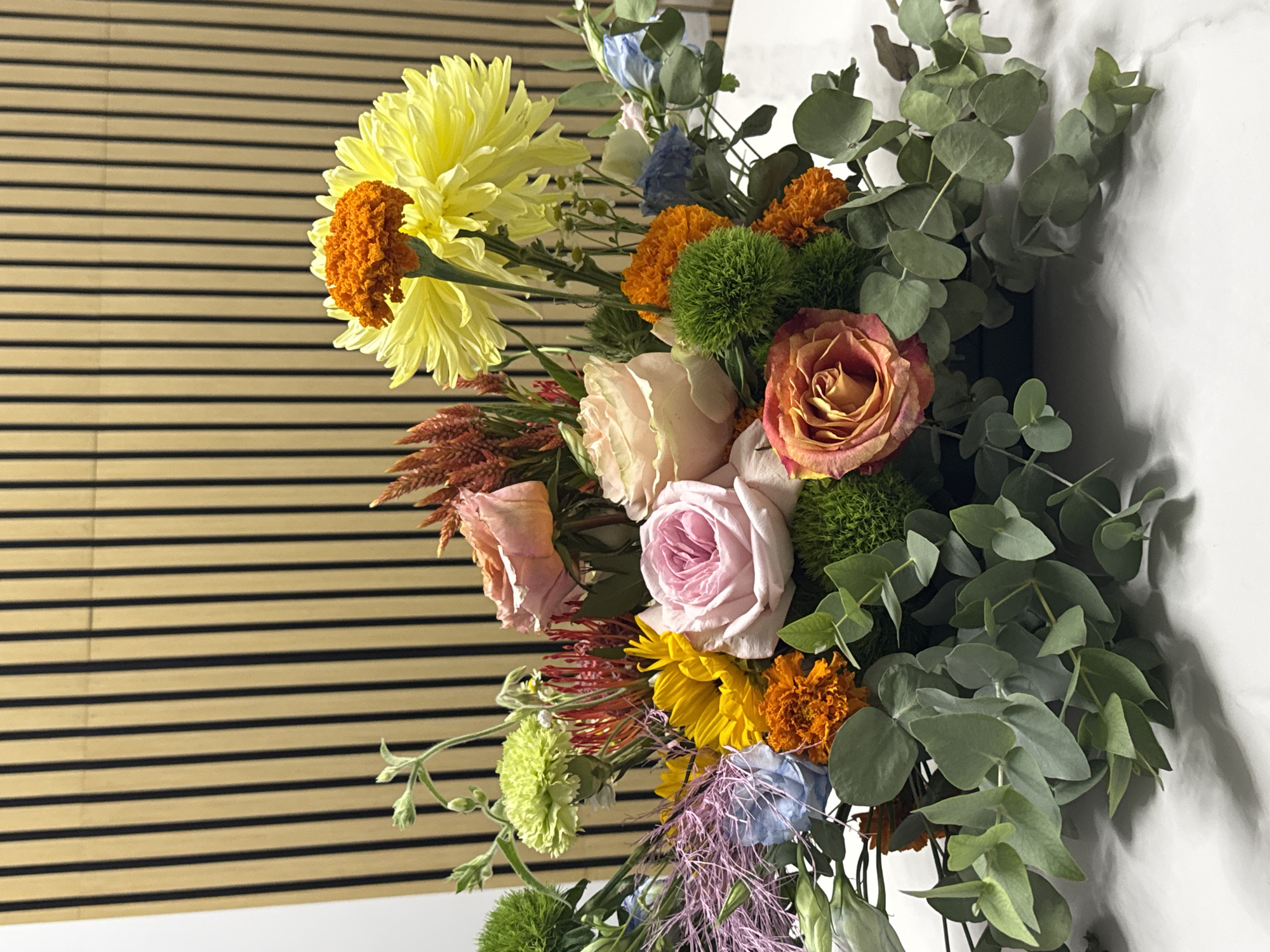 A colorful floral arrangement is displayed on a white marble surface in front of a vertical wooden slat wall. The arrangement features a variety of flowers including large yellow chrysanthemums with an orange center, pink and peach roses, orange marigolds, small green carnations, blue delphiniums, and wispy purple and red accents. Eucalyptus leaves form a lush base for the bouquet. The background wall consists of light-colored wooden slats with dark vertical lines, creating a ribbed texture. The lighting appears to be from above, casting subtle shadows on the marble surface. There are no people or animals present in the image. This scene does not provide any specific geographical markers to indicate its location within the city of Germasogeia Municipality, Cyprus.