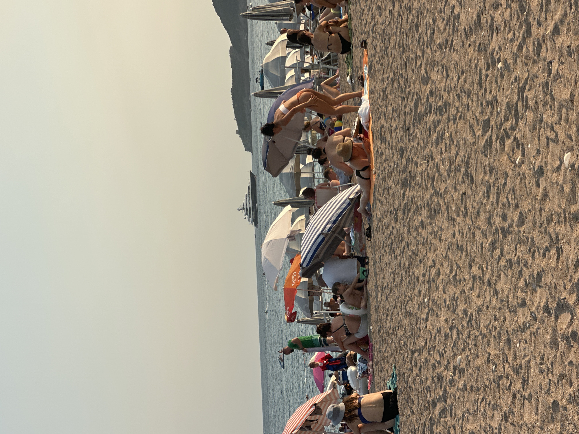 In Boreti, Montenegro, a crowded beach scene unfolds under a hazy sky. People are scattered across the sandy shore, many seated or lying under beach umbrellas, seeking shade from the sun. The sea stretches out in the background, with a large yacht visible in the distance, and smaller boats near the shore. Umbrellas of various colors and patterns dot the beach, providing shelter for beachgoers. Some individuals are wearing swimsuits, while others are clothed. The focus is on the leisure activities of people enjoying a day at the beach.