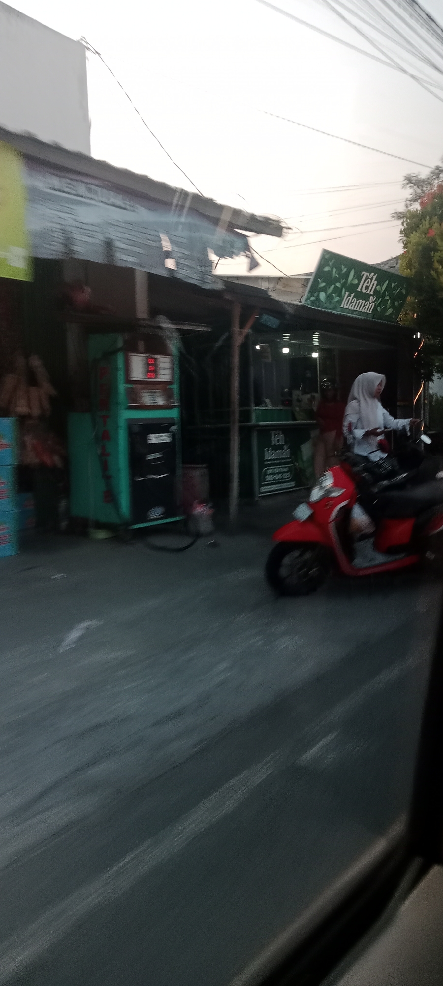 This image captures a street scene in Banyuraden, Indonesia, during what appears to be late afternoon or early evening, as indicated by the soft, diffused light and the clear sky. The foreground is dominated by a road with a slightly blurred texture, suggesting movement.
On the left side of the image, a teal-colored gasoline pump stands prominently. It has a digital display showing numerical values and appears to be operational. To the left of the pump, stacked blue boxes are visible. Behind the pump, a rustic structure with a corrugated metal roof is present, which seems to be part of a small shop or food stall.
Further to the right, a green sign with white text reading "Teh Idaman" (meaning "Desirable Tea") is mounted above another stall. This stall appears to be a beverage vendor, possibly selling tea, as suggested by the sign. Inside the stall, a person wearing a light-colored headscarf and a white shirt is seated on a red scooter. The scooter is positioned in front of the stall. The person on the scooter is looking towards the right, and their hands are on the handlebars. Another individual, possibly an employee of the stall, is partially visible inside the stall, wearing a dark top. The overall environment suggests a roadside commercial area.