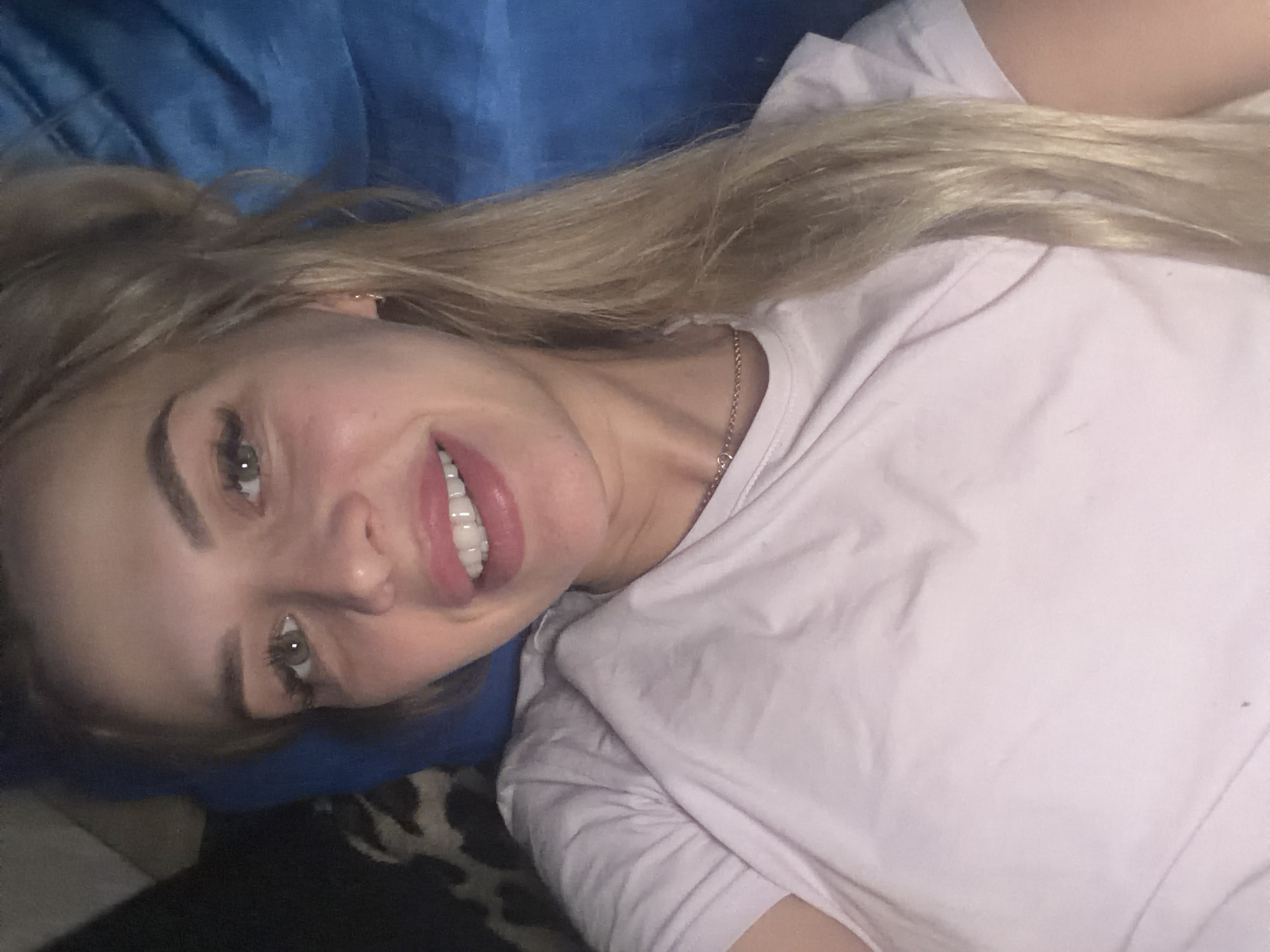 A young woman with blonde hair and bright makeup is lying down, looking into the camera. She is wearing a light pink t-shirt and a thin gold necklace. Her mouth is open, revealing white teeth. She is lying on a blue surface, possibly a bed or couch, with a dark patterned material visible in the background to the left. The lighting is dim and appears to be artificial. The image appears to be a selfie, taken from a close-up perspective. No specific location in Kyiv, Ukraine can be identified from the image.