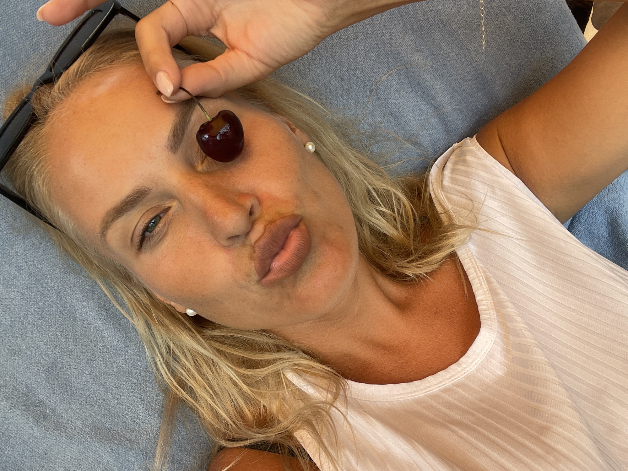 A woman with blonde hair and fair skin is lying on a blue textured surface, likely a towel or blanket. She is wearing a white ribbed tank top and pearl earrings. She is holding a cherry over her left eye, with a playful expression, puckering her lips. Black-rimmed glasses are resting on her forehead. The lighting suggests an outdoor setting, possibly during the day. The specific location in Baranovka, Russia, cannot be determined from the image itself.