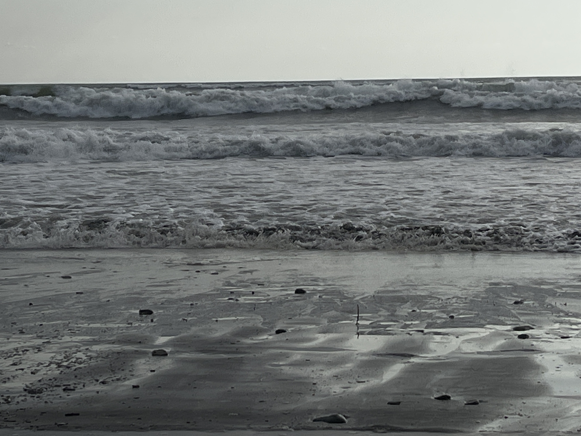 This media file displays a black and white image of waves crashing on a beach. The scene is dominated by the ocean with multiple layers of waves, some appearing larger and more forceful than others. The sky above is a pale, hazy gray, suggesting an overcast or diffused lighting condition, possibly indicating early morning or late afternoon.
The foreground features a wet sand beach with numerous small, dark pebbles scattered across its surface. The wet sand reflects the light, creating shimmering patterns and darker, damp areas. These reflections, along with the texture of the sand and pebbles, suggest the tide is either receding or has recently come in. There are no people or man-made objects visible in the frame, focusing the viewer's attention entirely on the natural elements of the sea and shore.
The overall mood of the image is calm and somewhat melancholic due to the monochromatic palette and the repetitive motion of the waves. The scene conveys the raw power and constant movement of the ocean. The location is identified as the Supsa community in Georgia. The time of day is not explicitly stated, but the soft, diffused light points towards a time outside of harsh midday sun. There is no visible text in the image.