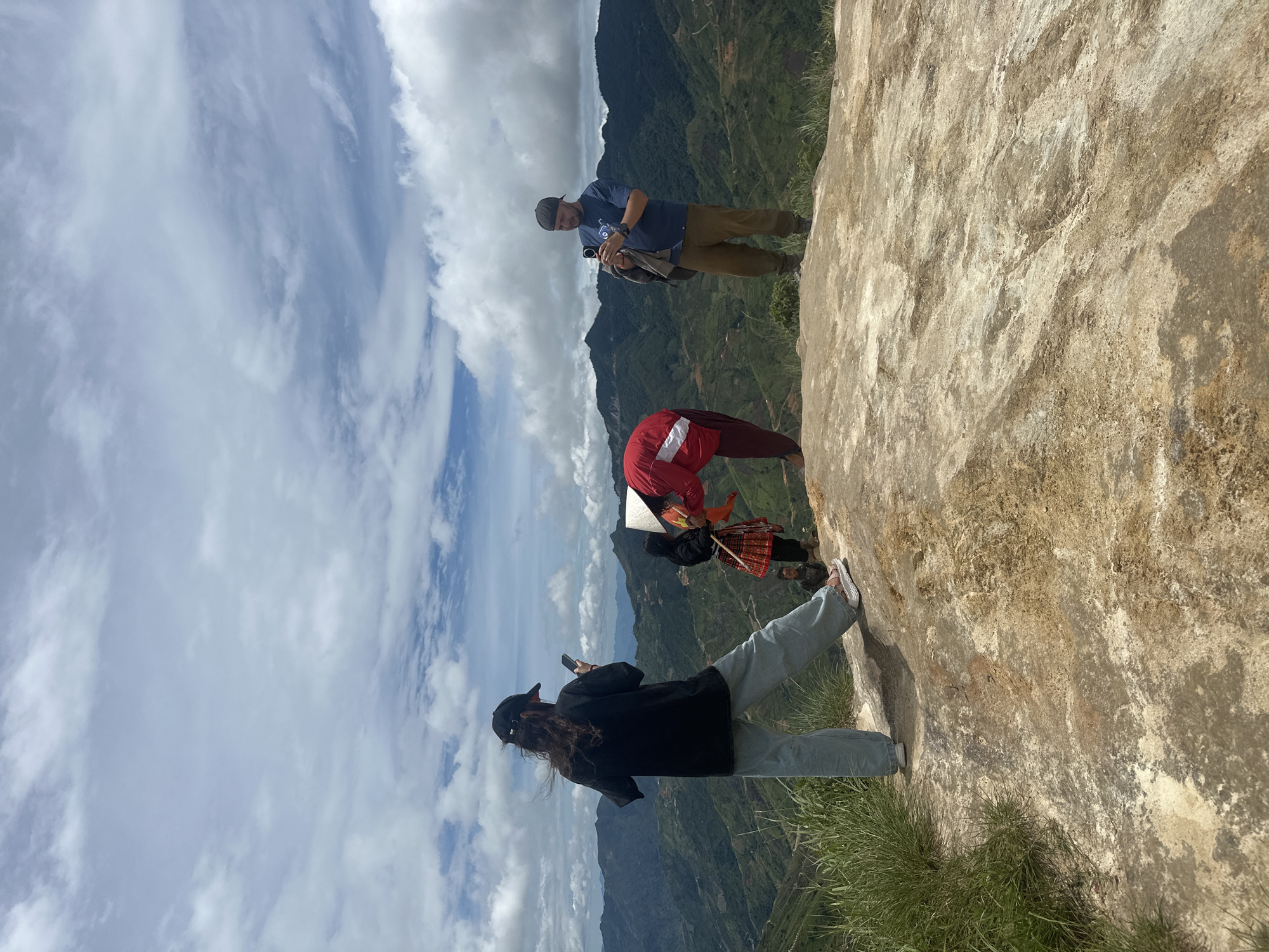 In Ta Xua, Vietnam, on a cloudy day, four individuals are positioned on a rocky outcrop overlooking a vast, mountainous landscape. The foreground is dominated by the textured, light-colored rock of the outcrop, with patches of dry grass growing at the edges. In the middle ground, a person wearing a black t-shirt and a black baseball cap is facing away from the camera, holding a smartphone. To their right, another person in a red jacket and conical hat is bending over, appearing to engage with someone or something on the ground. Further to the right, a person in a blue t-shirt and a baseball cap is standing and holding a smartphone, looking towards the left. In the background, the mountains of Ta Xua are covered in lush green vegetation, with visible terraced fields and winding roads. The sky is filled with a mix of white and grey clouds, suggesting a dynamic weather system.