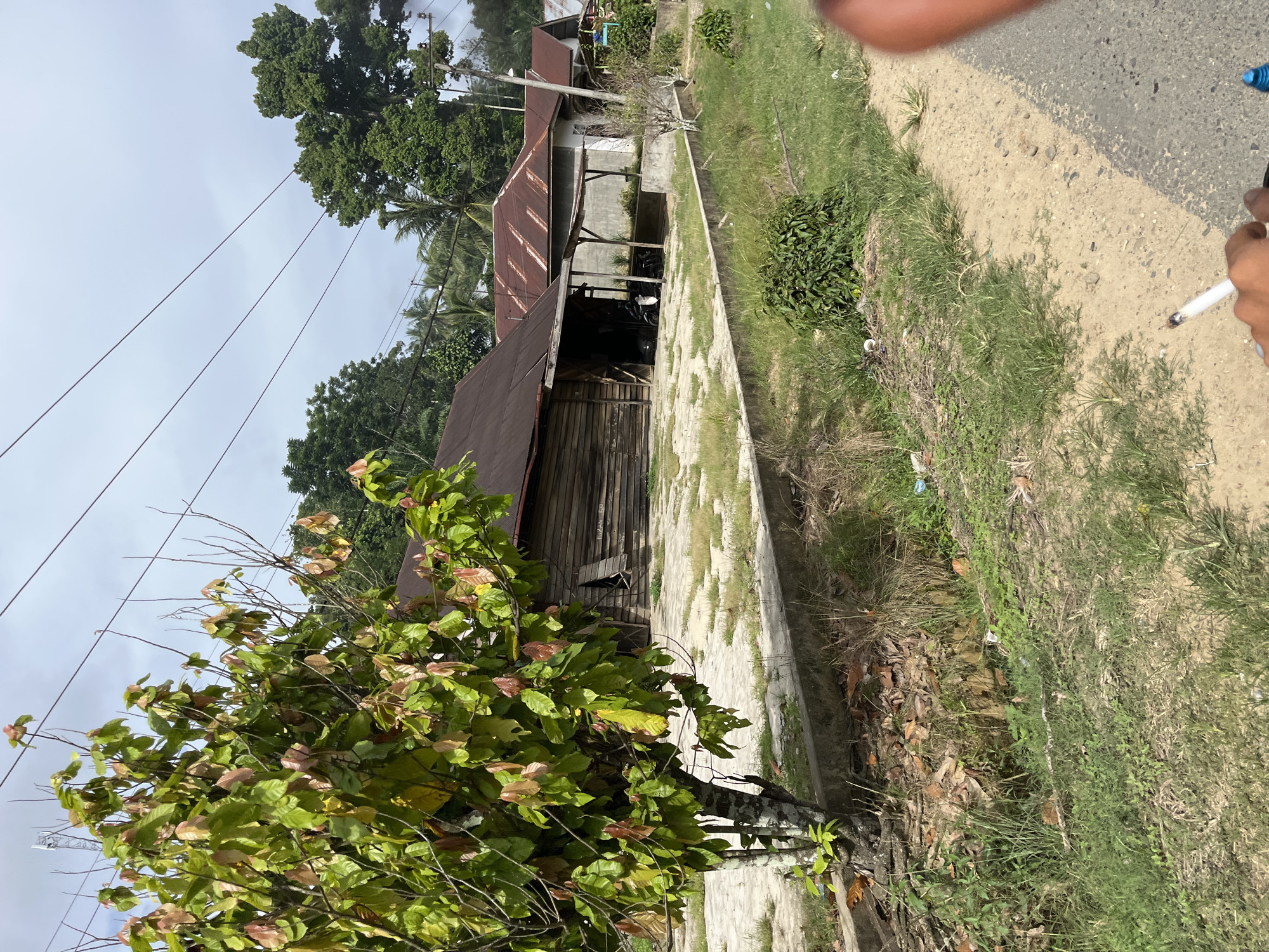 The image depicts an outdoor scene in Sukajadi, Indonesia. The perspective is from ground level, looking upwards and slightly to the side. In the foreground, a lush green tree with some reddish-brown leaves dominates the left side of the frame. Beyond the tree, a weathered wooden building with a rusty metal roof is visible. The building appears to be a simple structure, possibly a home or workshop, with an open-fronted section revealing dark interiors. A concrete path runs alongside the building, bordered by overgrown grass and vegetation.
To the right of the path, a dirt road or track is visible, with a hand holding a lit cigarette in the lower right corner of the image, suggesting a person is present. Another person's arm is visible in the upper right corner, with a blue object (possibly a lighter) nearby. The sky is overcast, indicating a cloudy day. The overall impression is a rural or semi-rural setting, likely during daylight hours. The presence of utility wires strung across the sky further suggests an inhabited area. The building's rustic appearance and the surrounding vegetation point towards a warm, possibly tropical climate.