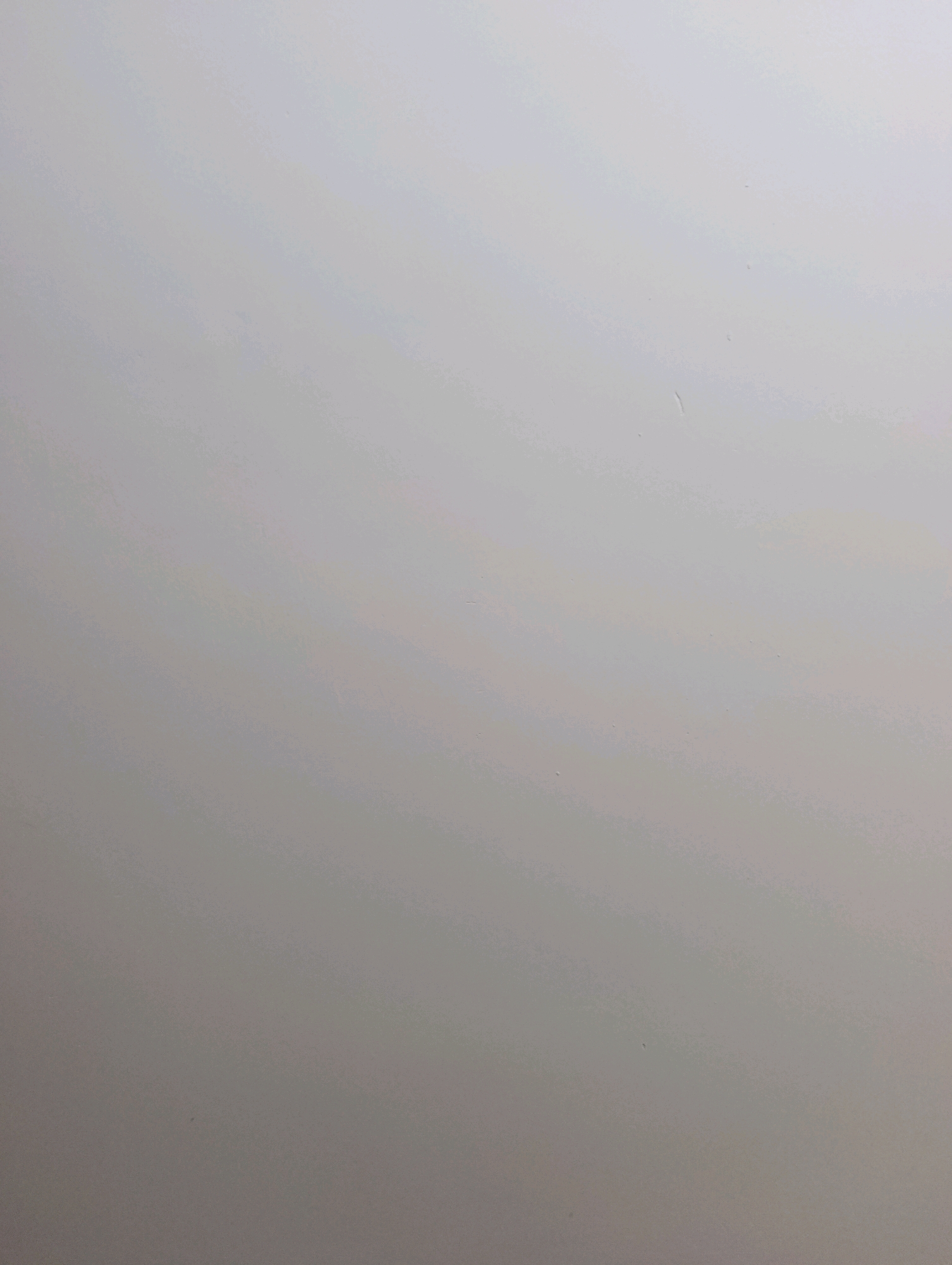 The image depicts a plain, light gray surface with a subtle gradient, appearing to be a wall or a similar flat material. There are a few minor imperfections or specks visible on the surface. The lighting suggests a diffused, ambient light source, creating a soft, even illumination without harsh shadows. The scene is devoid of any discernible objects, people, or activity, presenting a minimalist and abstract view of a surface. The location in Bhobtian, Pakistan, is not evident from the visual content of the image itself.