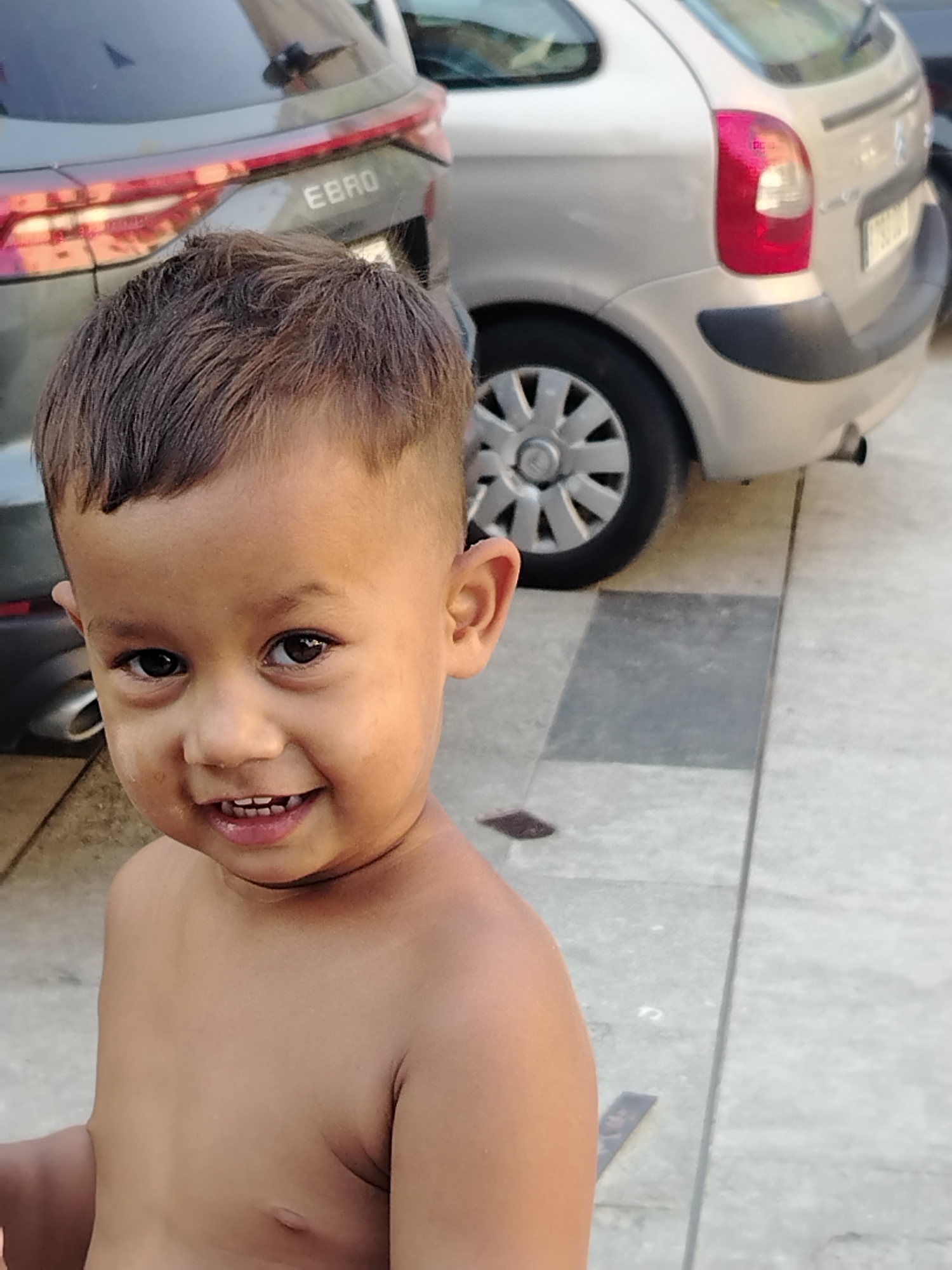 This is a close-up, medium-shot photograph of a young boy, likely of Hispanic descent, with dark hair and eyes. He is shirtless and looking directly at the camera with a wide, happy smile, revealing his teeth. The background is slightly blurred, showing the rear of two cars. To the left, a dark-colored SUV with "EBRO" visible on its tailgate. To the right, a silver hatchback. The cars are parked on a paved surface, possibly a street or parking lot. The lighting suggests it is daytime, with the sun casting subtle shadows. The overall emotion conveyed is joy and innocence. The setting appears to be outdoors in a casual, everyday environment. There is no obvious activity or event taking place other than the boy posing for the photo.