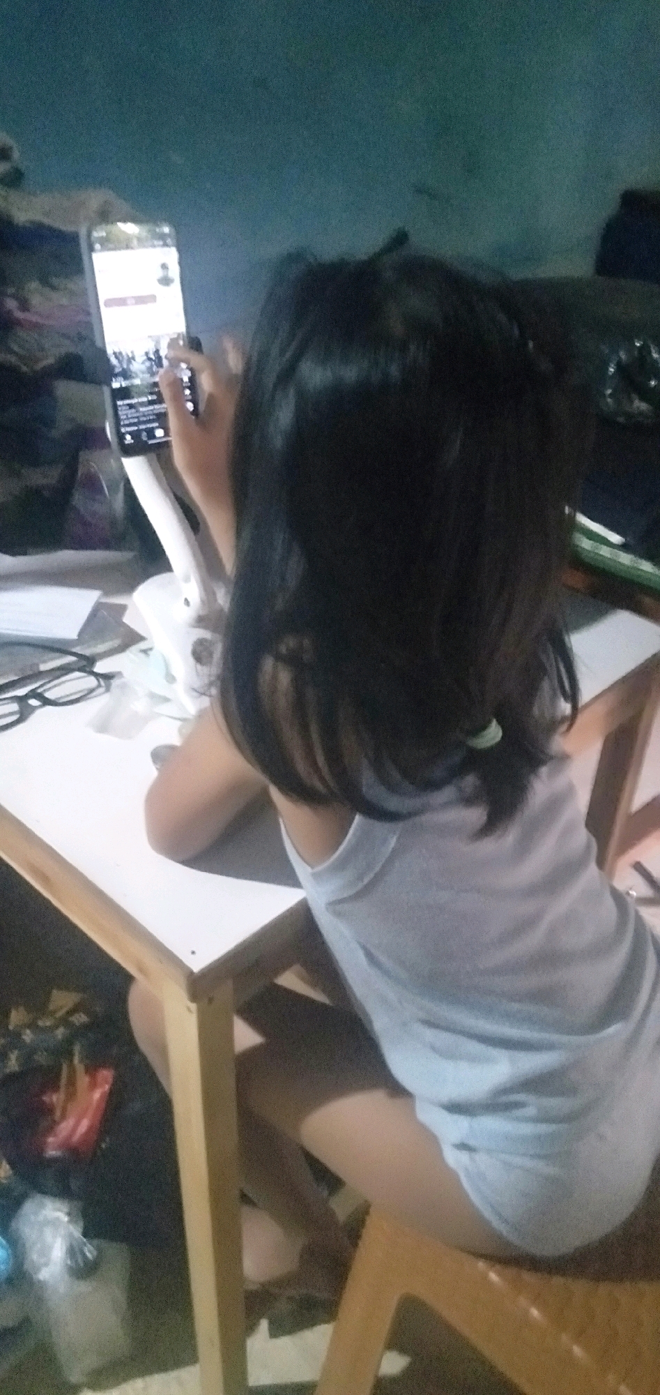The image shows a young person sitting at a white table and holding a smartphone mounted on a stand. The person is facing away from the camera and is wearing a light blue tank top. The background is a dark, mottled blue wall. On the table, there are a pair of eyeglasses, some papers, and other unidentifiable objects. The setting appears to be an indoor space, possibly a home, in Plemahan, Indonesia, given the provided context. The overall lighting is dim, suggesting it might be nighttime or a poorly lit room.
