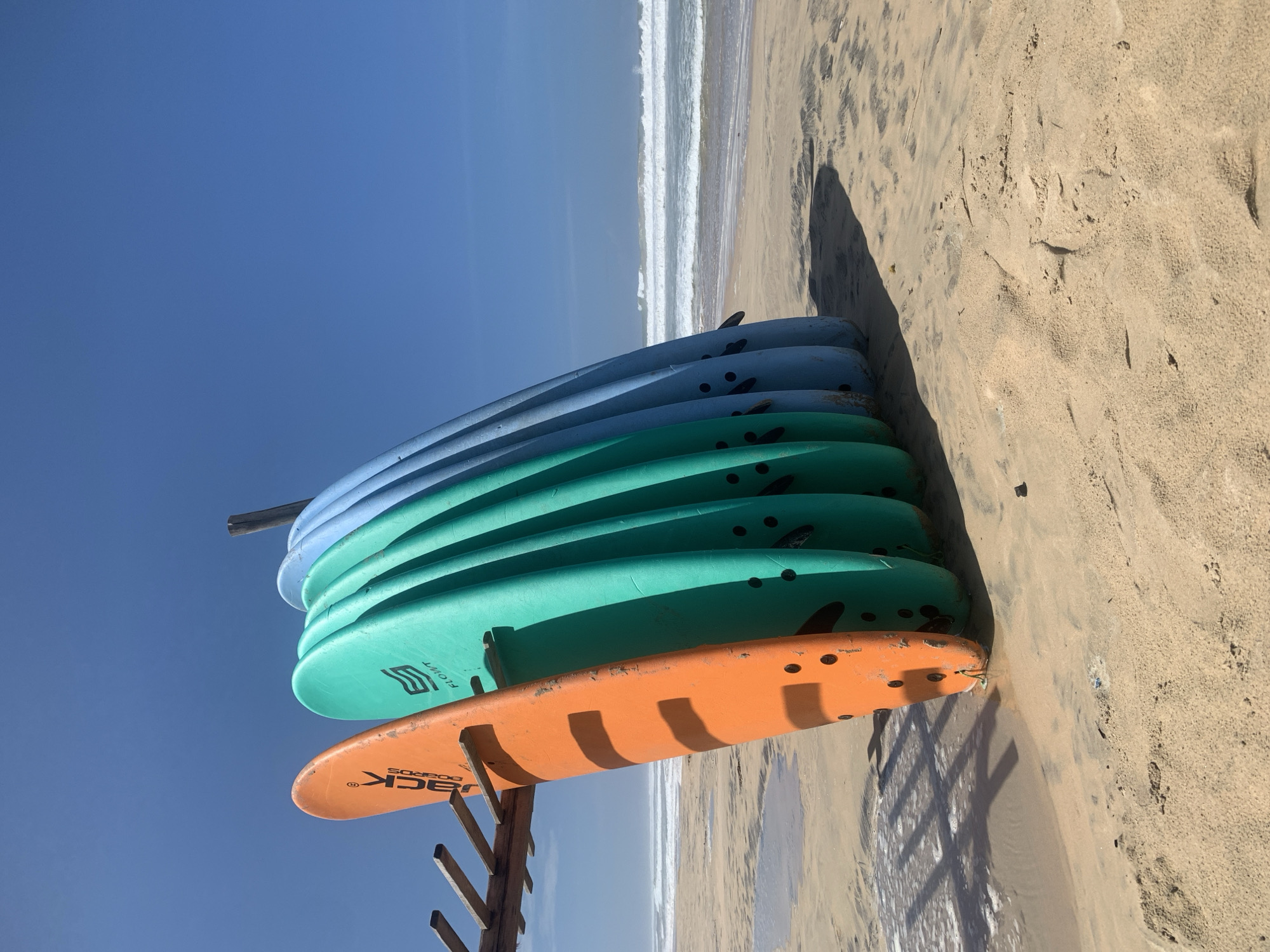 The image captures a stack of surfboards on a beach in Kathaluwa, Sri Lanka. The surfboards, varying in shades of blue, green, and orange, are neatly arranged on a wooden rack. The beach itself is sandy, with a visible shoreline where the ocean meets the land. The sky above is clear and blue. The scene is brightly lit, suggesting daytime.