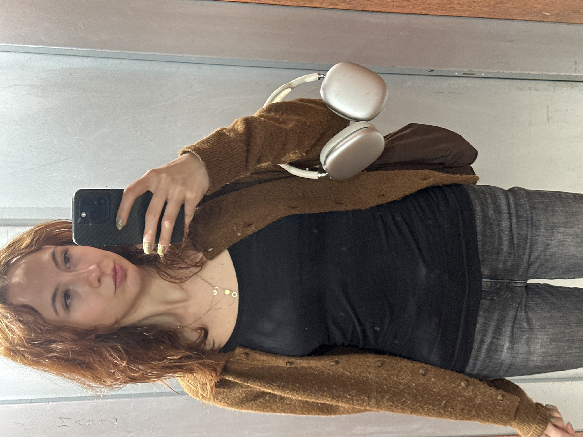 A young woman with long, reddish-brown, curly hair is taking a selfie in what appears to be a fitting room or elevator with light gray walls. She is holding a black smartphone with a textured case in her right hand. Her nails are painted with a light-colored polish. She is wearing a black top, a brown cardigan with buttons, and gray jeans. Around her neck is a gold necklace with circular pendants. On her left shoulder, there are white over-ear headphones. There is also a brown bag on her shoulder. The photo is taken in a slightly rotated orientation. The location in the city of Novi Sad, Serbia cannot be determined based on the image.