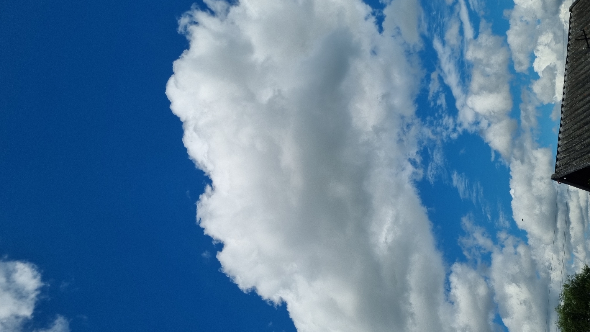 The image captures a clear blue sky filled with various white clouds. In the foreground, the corner of a dark, corrugated roof is visible, suggesting the presence of a building. A small dark object, possibly a bird or an insect, is seen in flight against the sky. The scene is set outdoors, likely in a rural or suburban area given the roof and the open sky. There are no people or animals visible in the image. The overall impression is one of a bright, cloudy day.