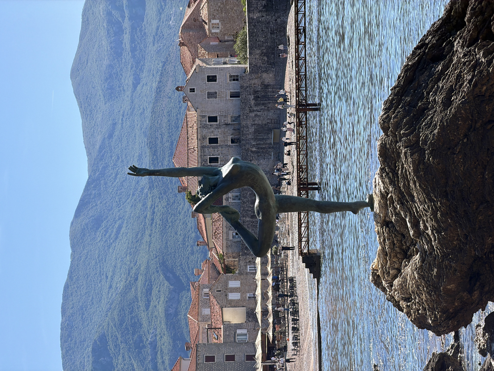 A bronze sculpture of a woman, known as the Budva Ballerina, stands on a rock jutting into the Adriatic Sea in Budva, Montenegro. The statue depicts the figure in a dance pose, with one leg extended and arms outstretched. Behind the statue, the architecture of Budva's old town is visible, including buildings with red tile roofs and stone facades. The scene is set against a backdrop of a mountain range. The water is calm, and the sky is clear. There are people scattered near the water's edge and on a walkway.