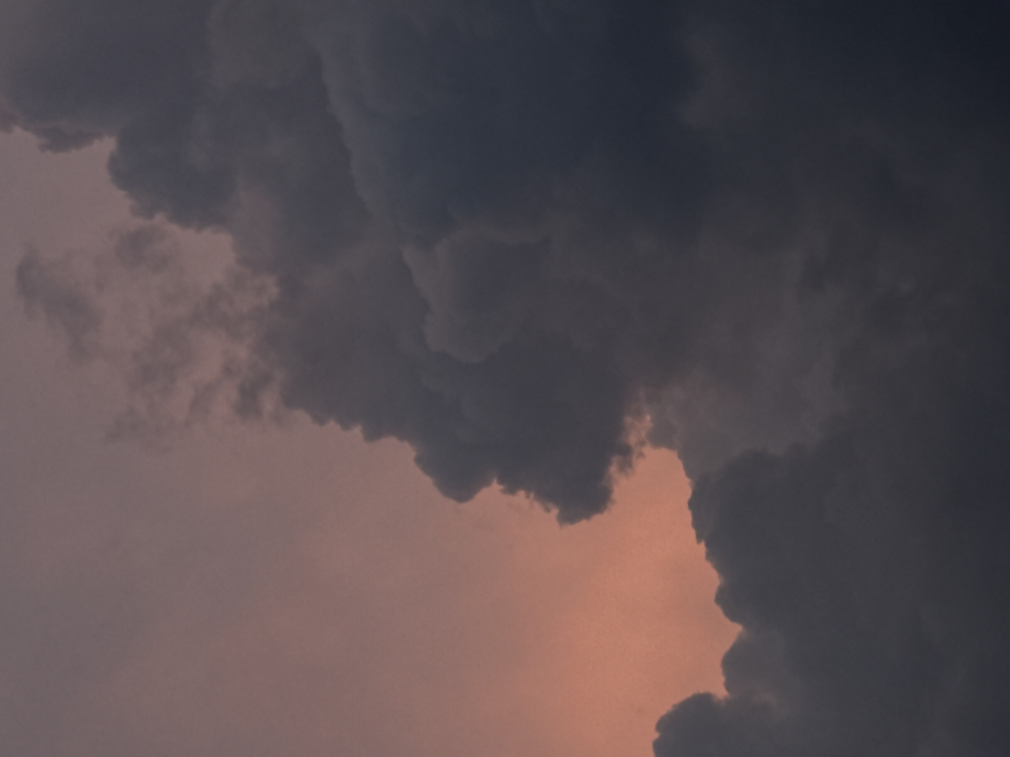 The image shows a dramatic sky filled with dark, ominous clouds against a soft, pinkish-orange sunset. The clouds are dense and voluminous, obscuring much of the sky. There is a stark contrast between the illuminated upper left portion of the sky and the shadowed, turbulent masses of clouds that dominate the right and lower parts of the frame. The texture of the clouds suggests movement and depth, with lighter areas hinting at reflected sunlight and darker areas indicating the thickness and density of the storm clouds. The overall mood is one of approaching weather and a sense of awe at the power of nature. The location is Savenki, Russia. There are no people, subjects, or objects visible in the image, only the sky.
