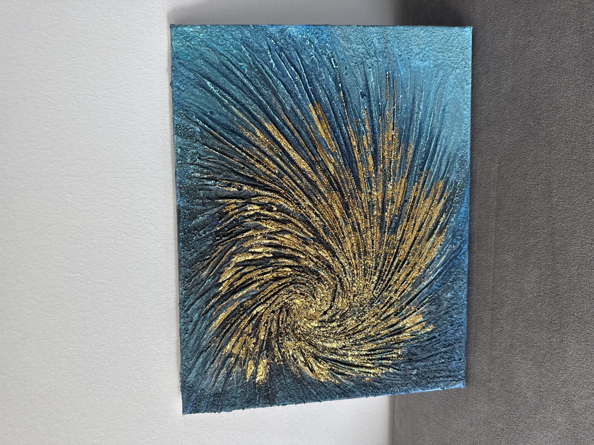 A textured painting with a swirling pattern of gold and dark teal is displayed against a white wall. The painting is placed on a grey surface, possibly a piece of furniture, and is angled slightly towards the viewer. The artwork features raised lines and a shimmering, metallic gold texture that radiates outward from a central point, creating a sense of movement. The background of the painting transitions from dark teal to a lighter, more vibrant teal on the right side. The texture of the painting appears rough and tactile. The white wall in the background has a subtle, uneven texture, and the grey surface below is a solid, flat color with a fabric-like grain. No people or animals are present in the image. The scene is static and appears to be an indoor setting.