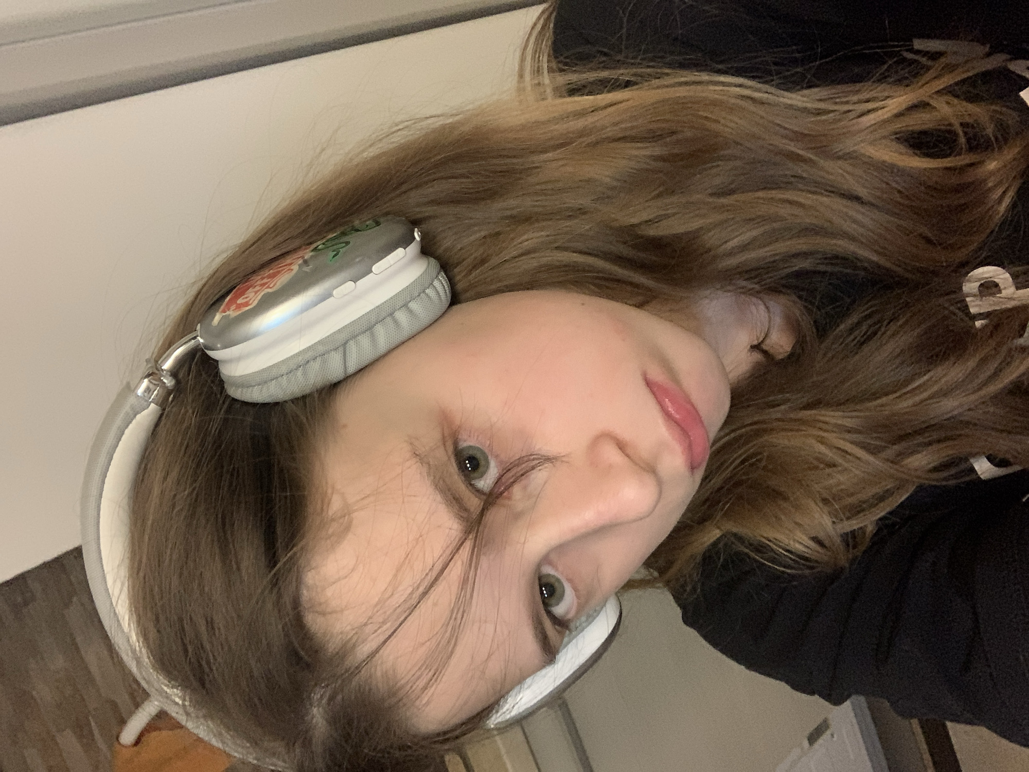 A young female with light brown hair is wearing grey headphones with a floral design on the ear cup. She is wearing a black top. The background is blurry and indistinct, showing a light-colored wall and part of a wooden surface. The image is rotated to the left.