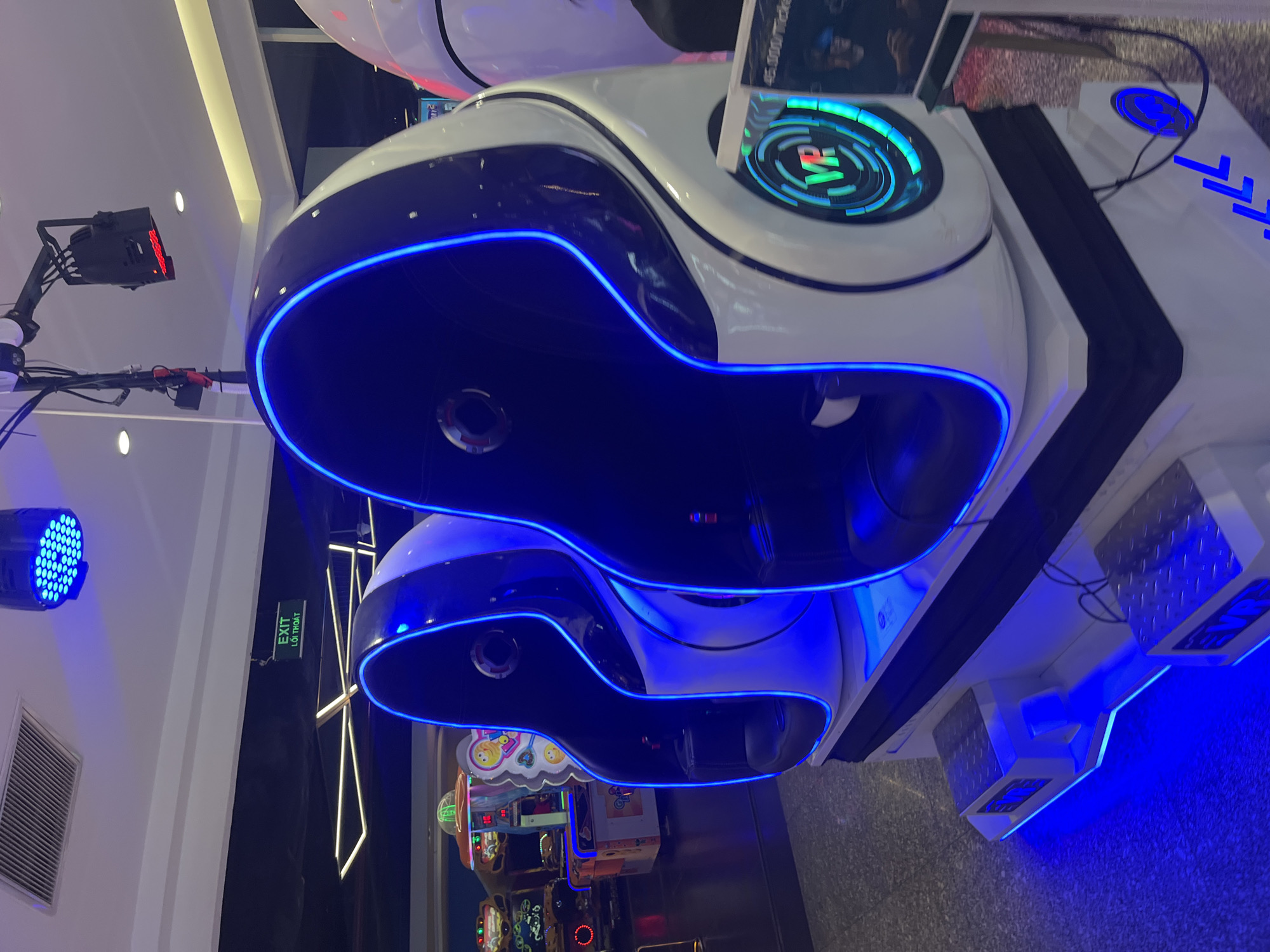 The image shows a pair of white and black virtual reality (VR) gaming pods, illuminated with blue neon lighting, located in an entertainment arcade setting. Arcade games are visible in the background, suggesting a location with recreational activities. The specific location is Nha Trang Ward, Vietnam. A black light and exit sign are mounted on the wall to the left of the VR pods.