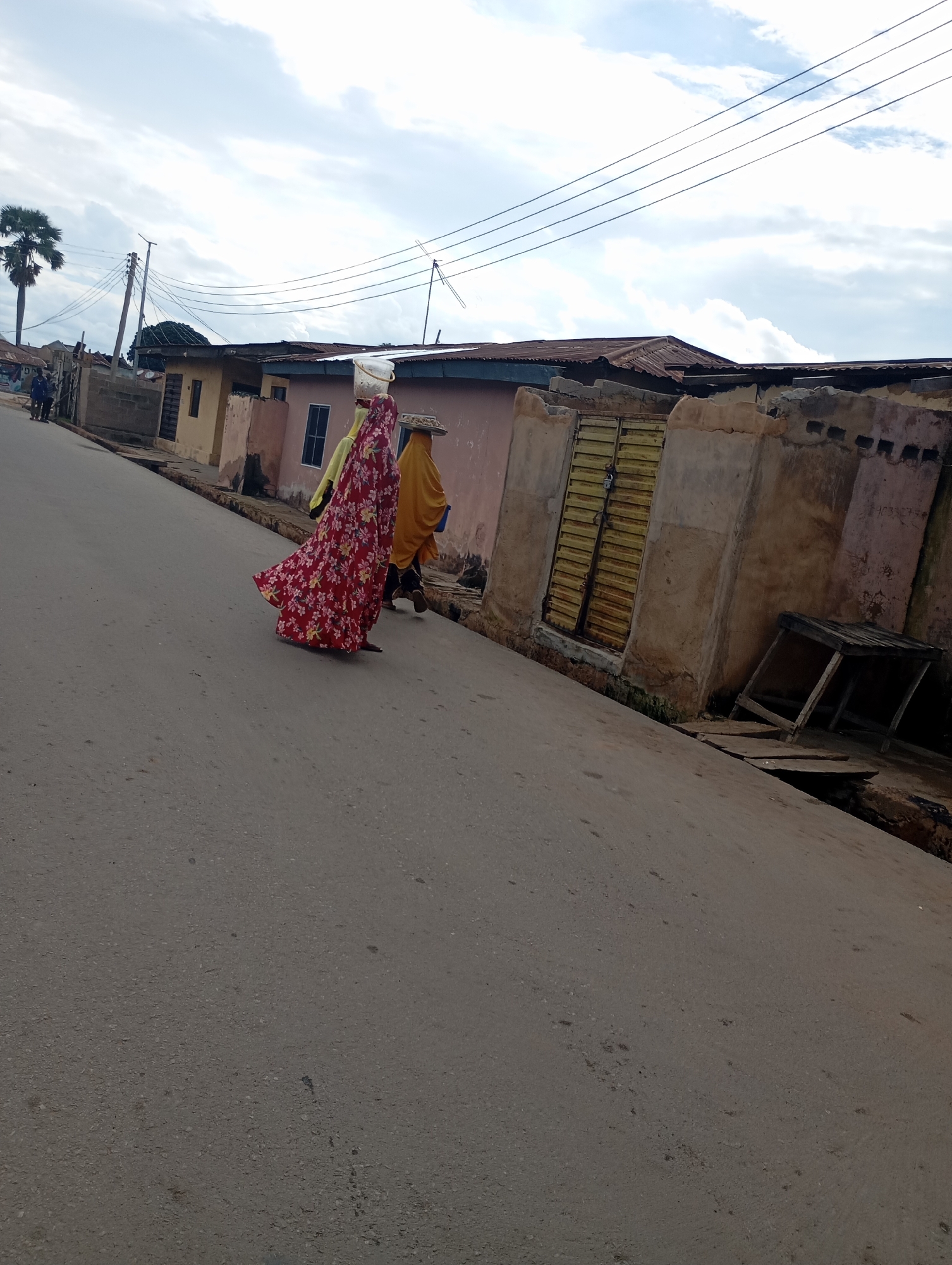 In Shendam, Nigeria, two individuals walk down a paved road. The person in front wears a vibrant red dress with a floral pattern and carries a white bucket on their head. Behind them, another person is dressed in yellow and appears to be carrying a flat object on their head. They are walking past a row of buildings with rustic, weathered walls and corrugated metal roofs. A yellow metal gate, padlocked, is visible on one of the buildings. A wooden table structure stands near the gate. Overhead, power lines stretch across a partly cloudy sky. A palm tree can be seen in the distance. The road surface is asphalt.