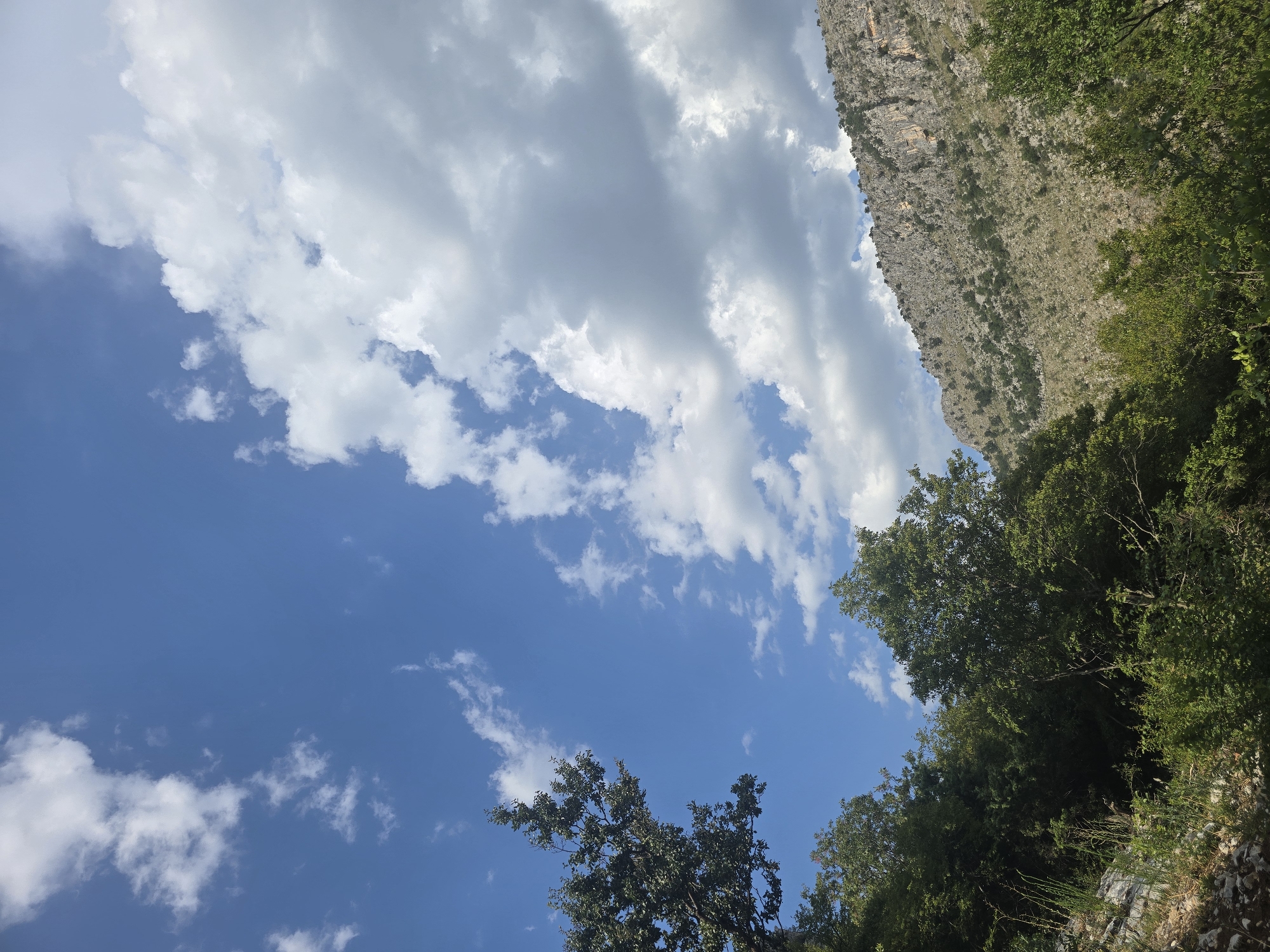 This is a vertical, eye-level, full-shot photograph of a mountainous landscape under a partly cloudy sky. The sky is a vibrant blue with scattered white cumulus clouds. In the foreground, lush green trees and foliage are visible, with some rocky terrain to the left. The middle ground features a steep, rocky mountainside dotted with sparse vegetation. The scene appears to be a natural, outdoor environment. There are no people or animals present in the image. The lighting suggests it is daytime, with the sun likely positioned to the right of the frame, casting some shadows. The location is identified as the city of Podgorica, Montenegro.