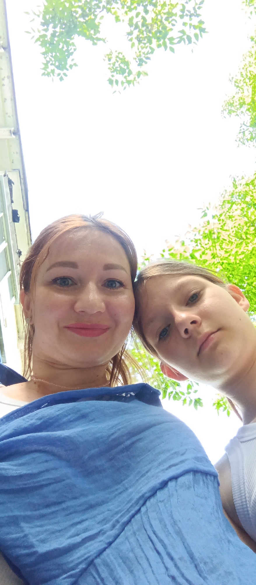 Two individuals, a woman and a child, are depicted in the image, taken from a low-angle perspective. The woman has light skin, red hair, blue eyes, and pink lipstick. She is wearing a light blue garment. The child, positioned slightly behind and to the right of the woman, has light skin, light brown hair, and blue eyes. The background reveals partial views of foliage and a building exterior. The location is described as a city named Unknown in Unknown.