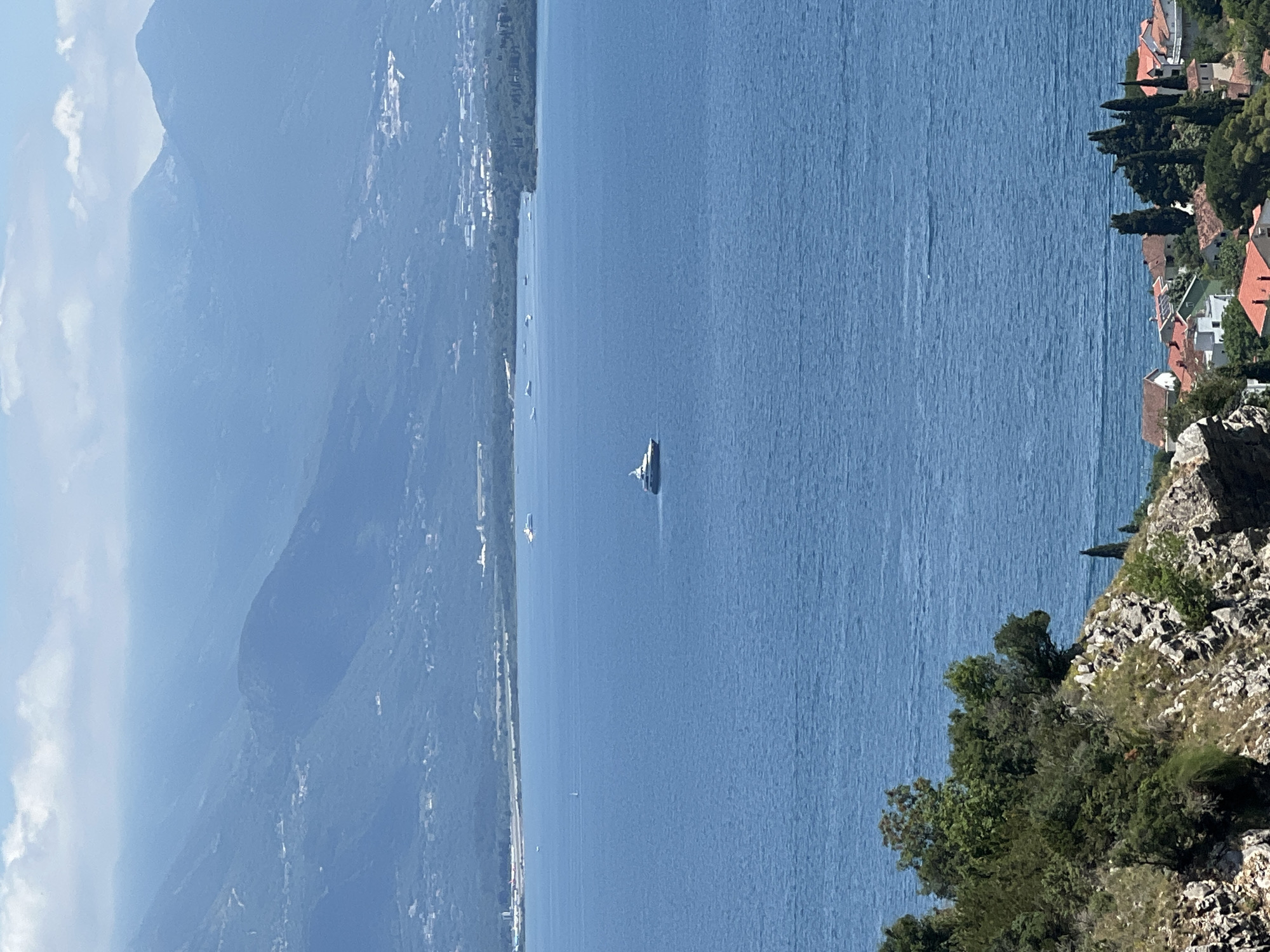 The image depicts a serene coastal landscape, possibly in Baošići, Montenegro. A yacht is visible in the calm, blue waters, with a backdrop of mountains and coastal settlements. The foreground shows rocky terrain with vegetation, suggesting a viewpoint from an elevated position. Houses with red-tiled roofs dot the landscape near the coast on the right side of the frame. The sky is clear, indicating bright daylight.