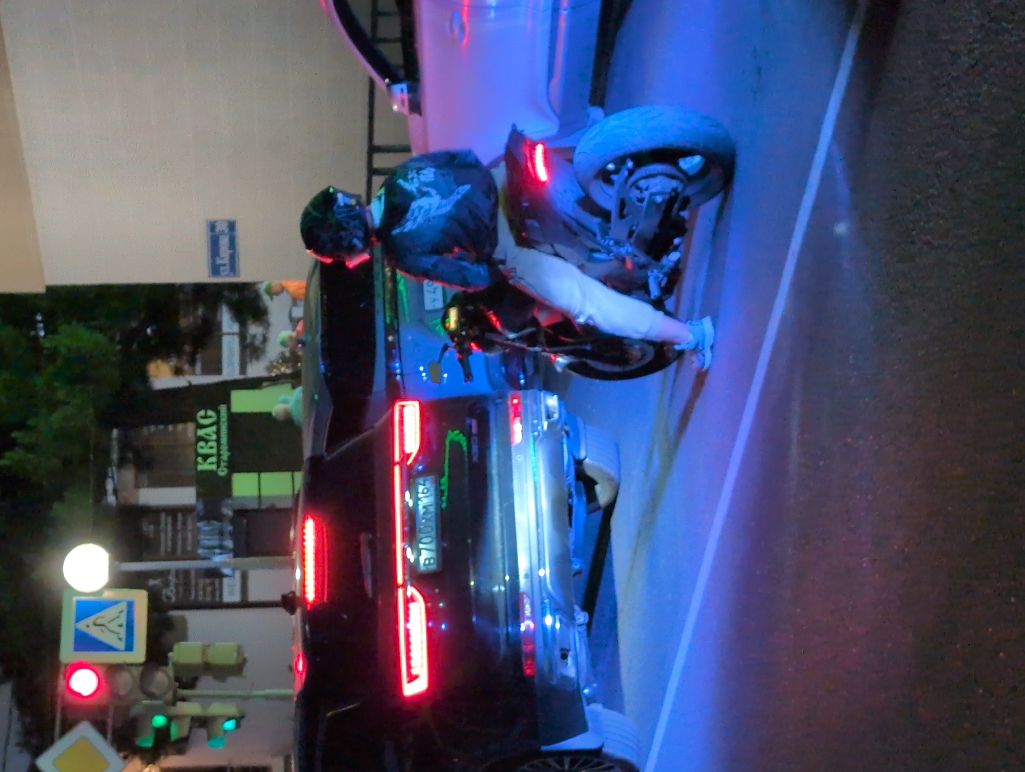 The image depicts a street scene at night, likely in Gelendzhik, Russia, as indicated by street signage and vehicle license plates. In the foreground, a person is seated on a motorcycle, facing away from the camera, wearing a helmet with a reflective visor, a dark jacket with a winged design, and light-colored pants. The motorcycle has a red taillight illuminated. To the left of the motorcycle, a black SUV is parked, its brake lights and taillights glowing red. The SUV's license plate is visible, reading "B700XM 164". Across the street, a white car is parked. Traffic lights are visible on the left side of the image, with a red light illuminated. A pedestrian crossing sign is also present. In the background, a building with signage that reads "КВАС" (Kvass, a fermented beverage) is visible, along with a street sign that appears to read "ул. Карина 30" (Karina St. 30). The scene is illuminated by artificial light, creating a mixture of colors, including blue and red hues.