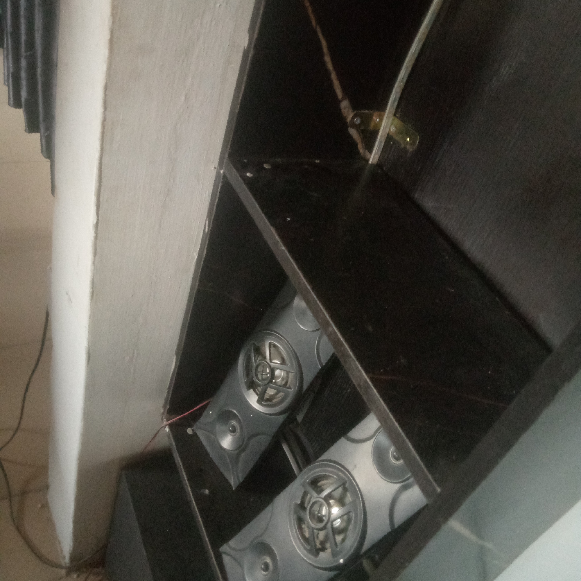 The image shows two gray speakers on a shelf within a dark wooden cabinet. The speakers are rectangular with circular speaker grilles and smaller circular accents. One speaker is positioned slightly in front of the other. Red and black wires are connected to the speakers. To the left of the cabinet is a light-colored wall with a ribbed texture at the top. The scene is indoors, likely in a room with ambient lighting. The overall impression is one of a home entertainment setup.