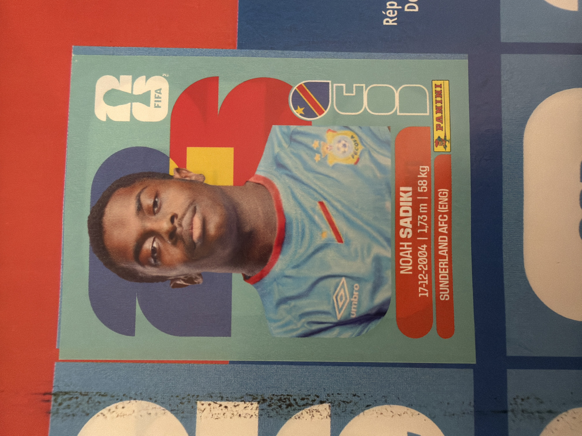 The image is a close-up of a football sticker featuring Noah Sadiki. He is wearing a blue jersey with a red and yellow trim and a crest on the left chest. Text below his image reads "NOAH SADIKI", his birthdate "17-12-2004", his height "1,73 m", and his weight "58 kg". Below that, it states "SUNDERLAND AFC (ENG)". A small Panini logo is visible to the right. The sticker is positioned on a red and blue background, suggesting it is part of a larger collection. There is no information in the image or provided crops that indicates the location is in Zlatitrap, Bulgaria.