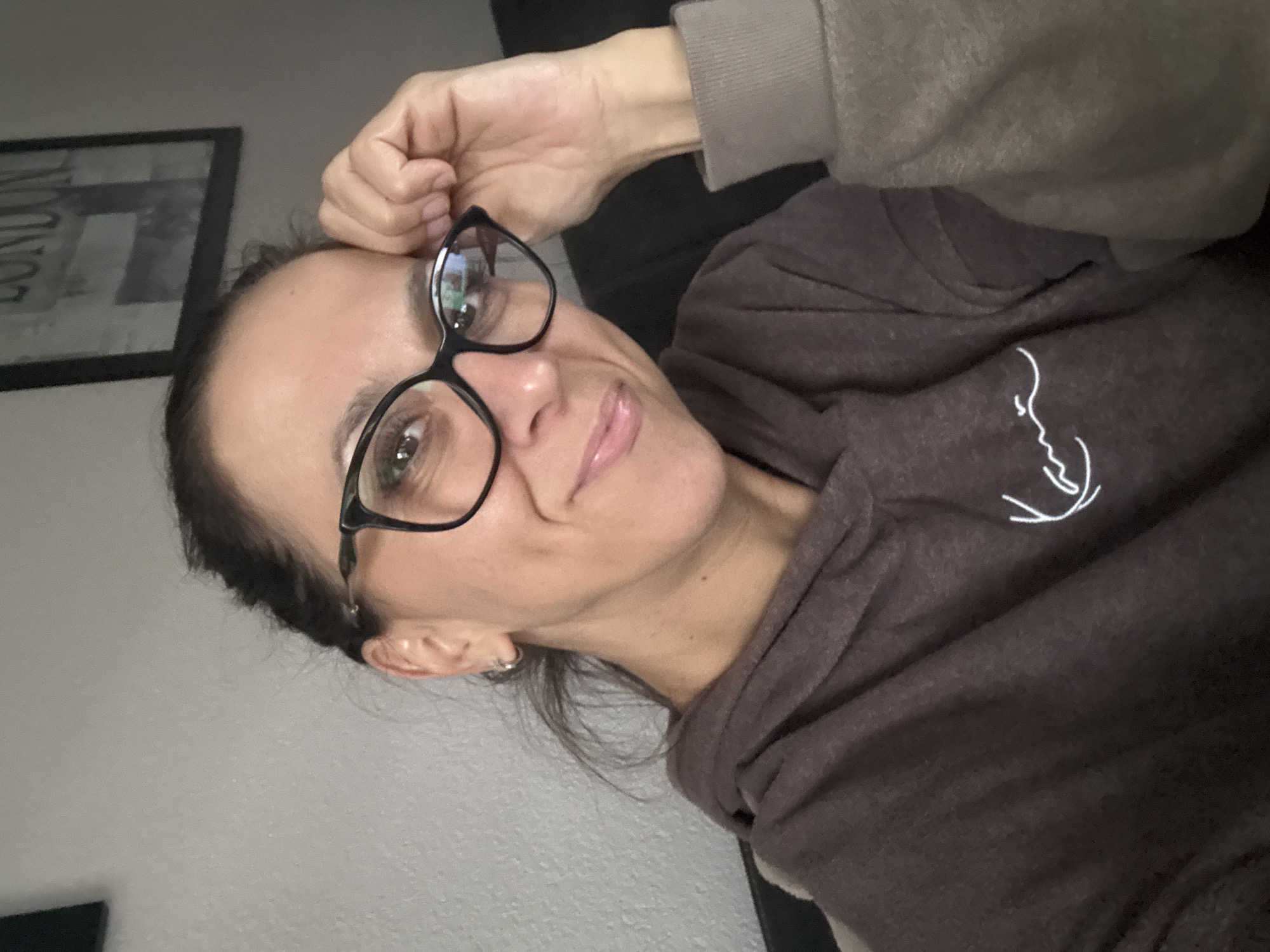 A woman with dark hair pulled back and wearing glasses is looking at the camera, with her hand touching her temple. She is wearing a brown hoodie. The background is a light gray wall with a framed picture on it. There is also a dark surface behind her that appears to be a couch. There is no indication of the city of Marum, Netherlands in the image.