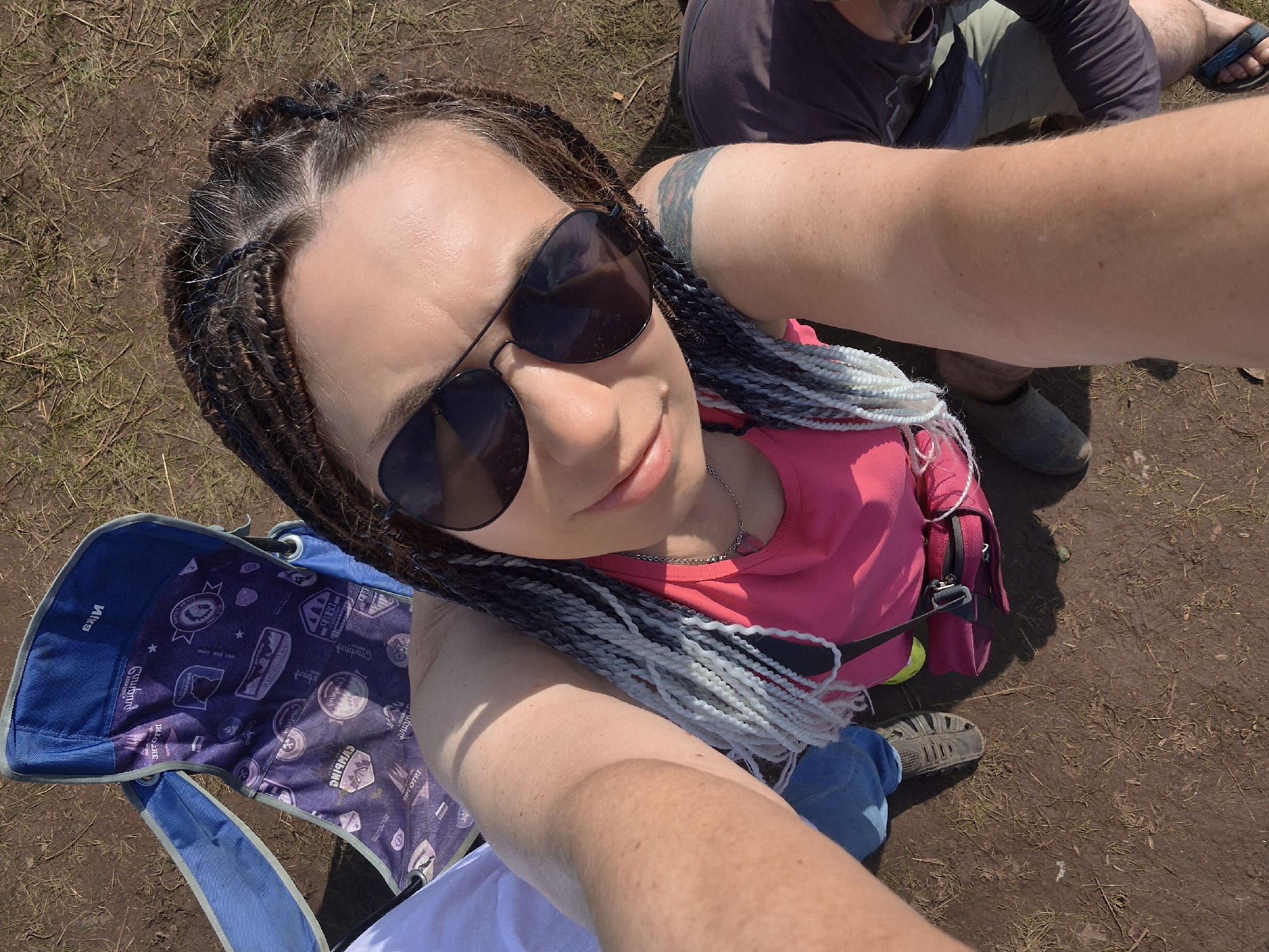 A high-angle, medium shot captures a woman with braided hair wearing sunglasses and a pink tank top, taking a selfie outdoors. She is seated on a blue camping chair with a purple, patterned seat. Her right arm is extended towards the camera. To her left, a person wearing a white shirt is partially visible. To her right, another person wearing a dark shirt and olive-green pants is visible, with their legs and feet in view. The ground is a mixture of dirt and dry grass. The overall scene suggests a casual outdoor gathering or event.