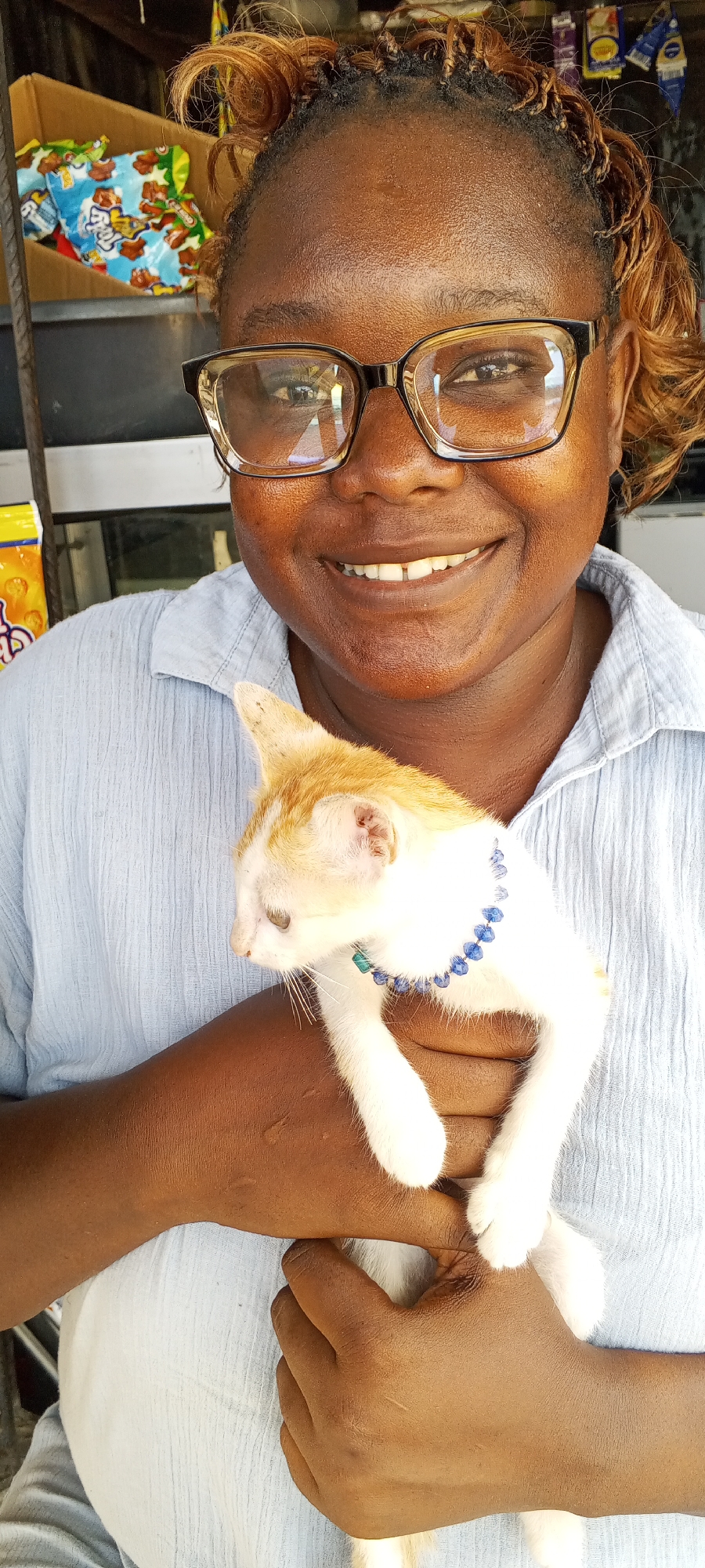 A smiling woman wearing glasses and a light blue button-down shirt holds a white and orange kitten with a blue beaded collar. The background includes a shelf with food items, suggesting a shop or market. The location is Lagos, Nigeria.
